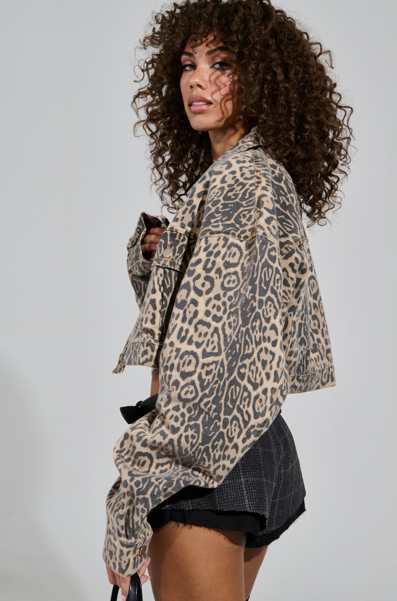 A person with curly hair poses in a cropped NO CHEATING PRINTED DENIM JACKET paired with dark shorts while holding sunglasses, set against a plain, light-colored background.