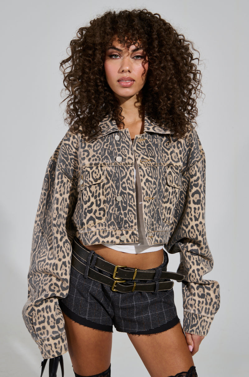 A person with curly hair is wearing the NO CHEATING PRINTED DENIM JACKET and black plaid shorts featuring double belts. The cropped silhouette accentuates their style as they look confidently at the camera.