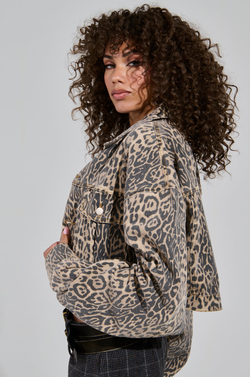 A person with curly hair is wearing the NO CHEATING PRINTED DENIM JACKET in a cropped leopard-print style and is looking over their shoulder against a plain background.