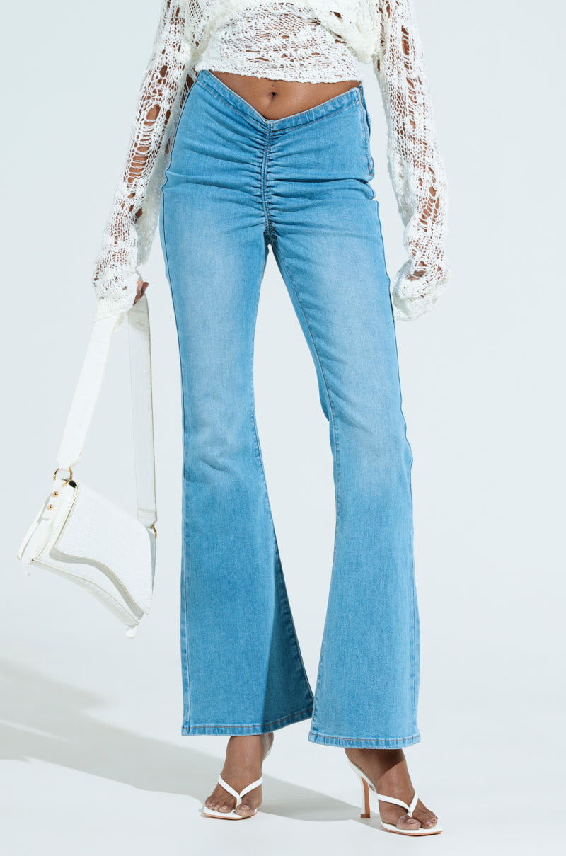 A person in CINCHED UP DENIM PANTS IN MEDIUM WASH and a white semi-transparent crochet long-sleeve top holds a white handbag. They're wearing white heeled sandals, and the jeans feature gathered detailing at the front.
