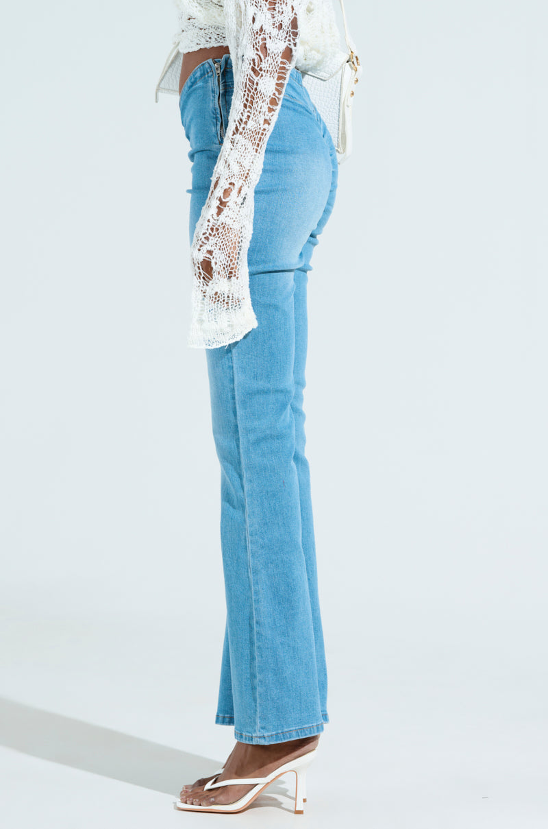 Someone is wearing CINCHED UP DENIM PANTS IN MEDIUM WASH with a white lace top and white high-heeled sandals, standing sideways against a plain, light background.