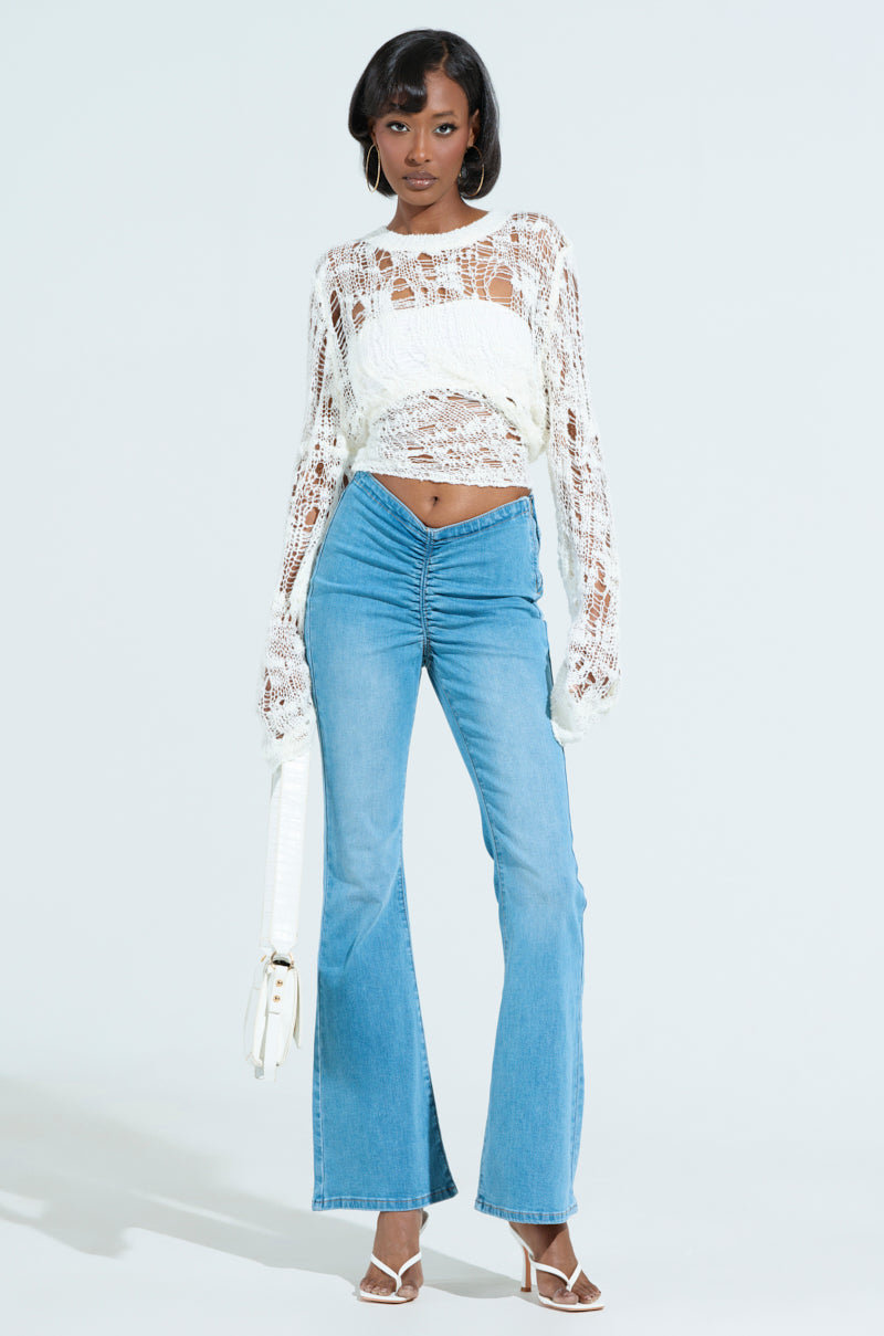 Dressed in a white, long-sleeve crochet top and CINCHED UP DENIM PANTS IN MEDIUM WASH with a flared leg, the person stands confidently against a light background. They're accessorized with hoop earrings, open-toed heels, and carrying a white handbag.