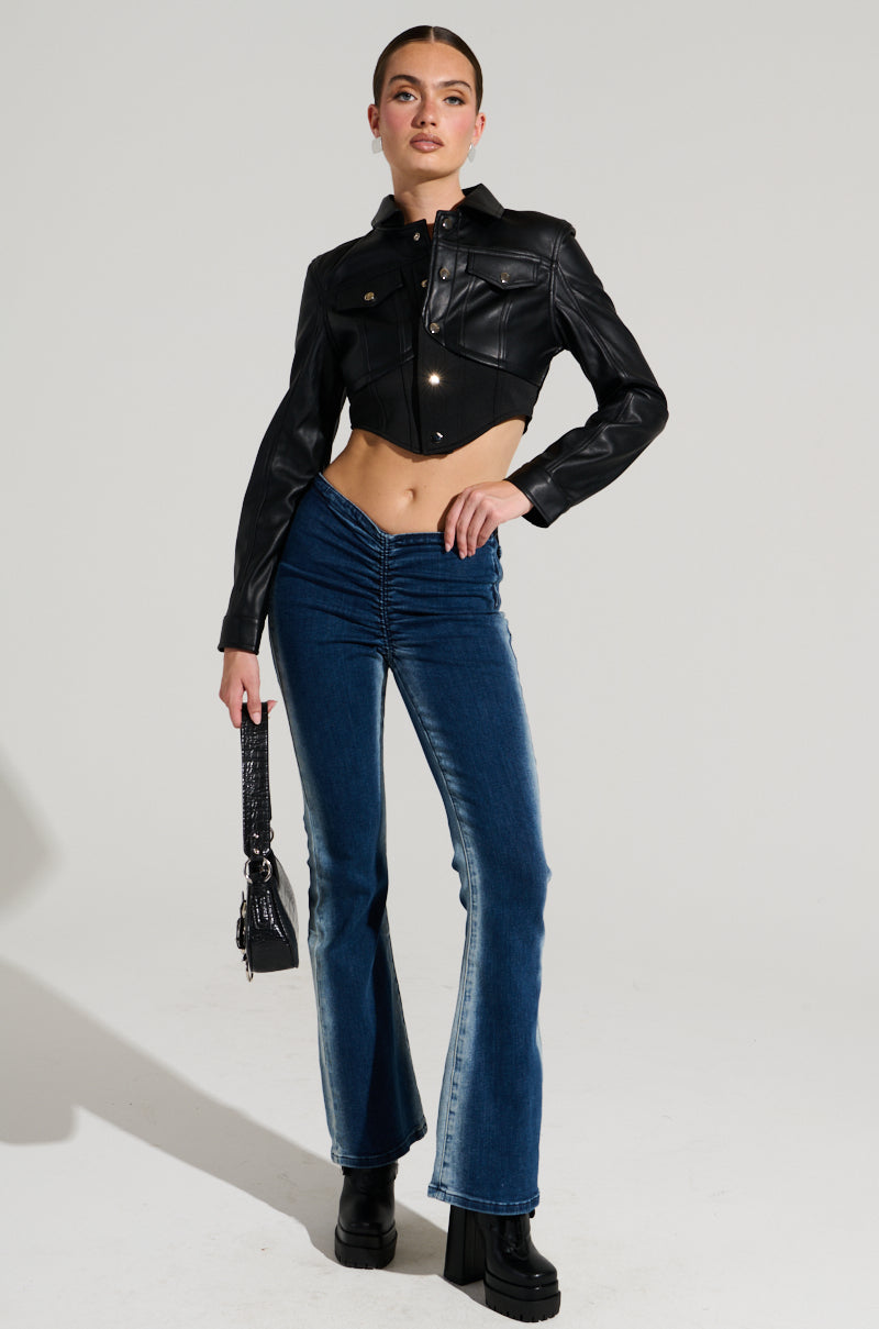 A person with light skin and long hair tied back is wearing a black leather cropped jacket, Cinched Up Denim Pant in Medium Blue Denim with ruched seaming at the front, and black platform boots. They are holding a small black handbag and standing in a confident pose.