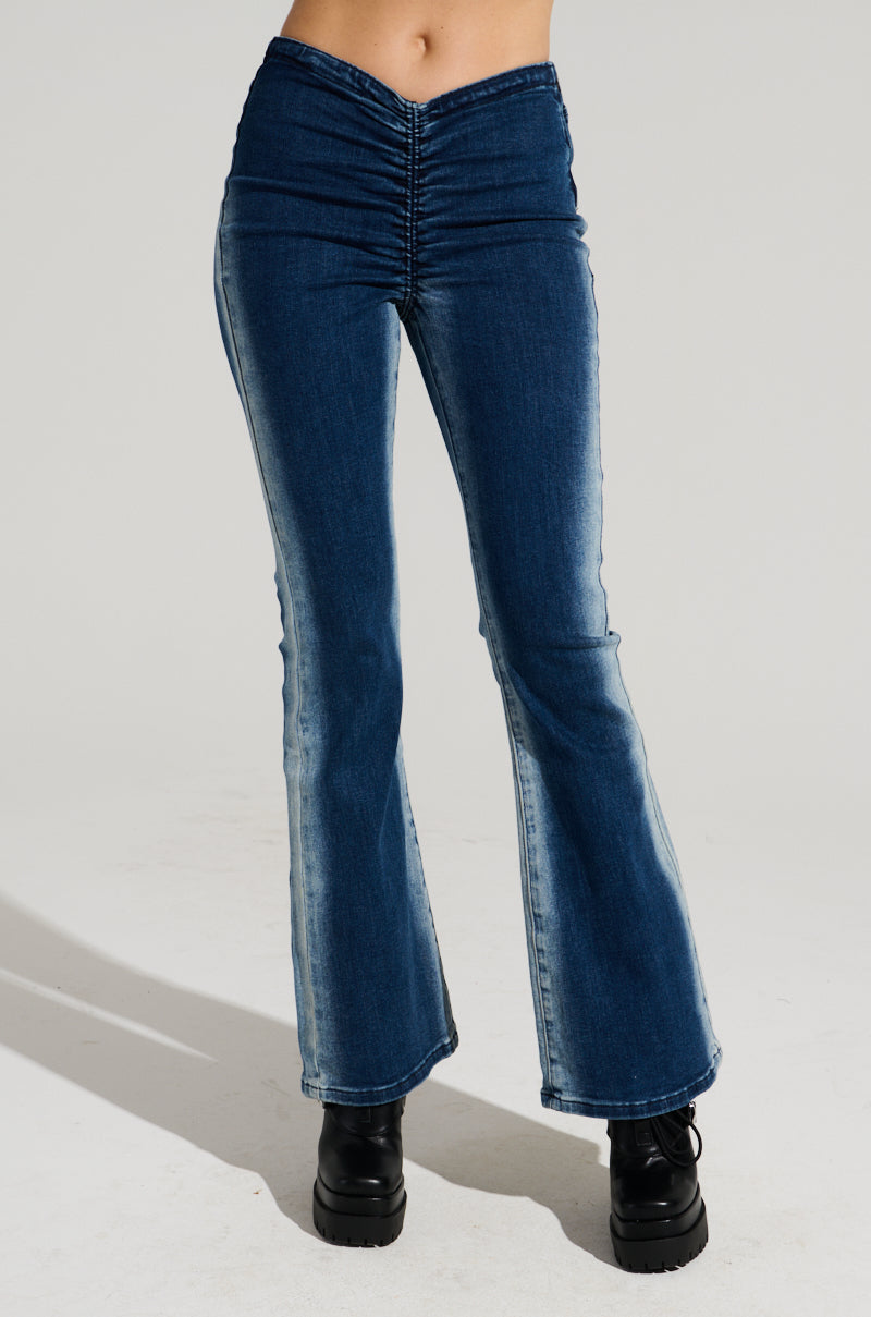 A person wearing high-waisted, medium blue Cinched Up Denim Pants with ruched seaming at the waist and black platform boots. The jeans have a slight fade on the thighs and knees. The background is plain and light gray.