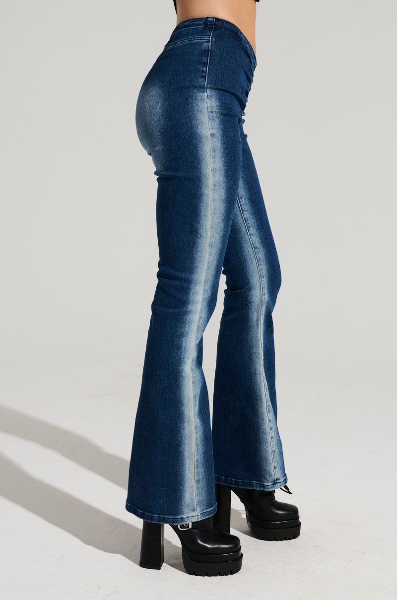 A person is modeling the CINCHED UP DENIM PANT IN MEDIUM BLUE DENIM, featuring a high-rise v-shaped waist and a flared cut with a faded stripe down the side. They are also wearing black platform boots. The background is plain and light-colored.