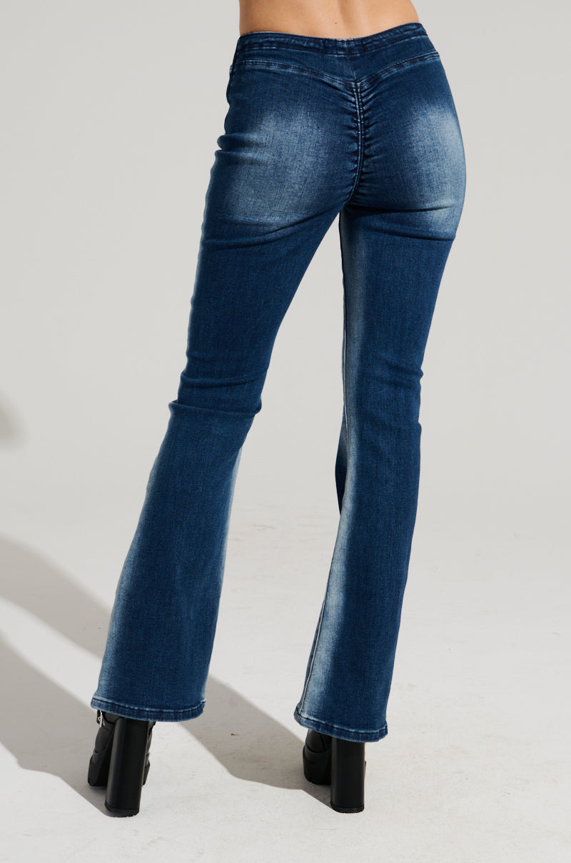 A person is wearing medium blue, Cinched Up Denim Pant jeans with a high rise v-shaped waist and slight fading along the thighs and back. The fitted design features flared bottoms and ruched seaming. Standing on a neutral background, their black shoes with thick heels are partially visible.