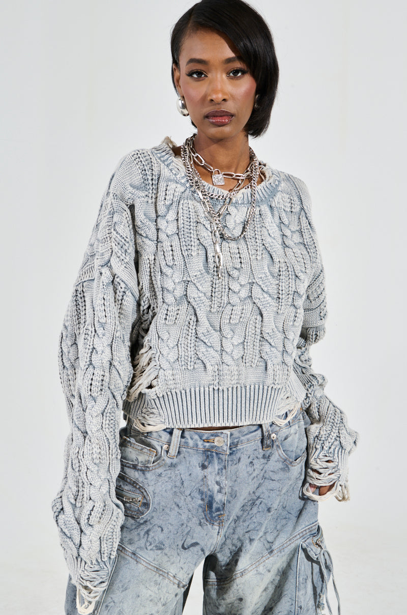 A person poses confidently in an AKIRA Label TALK TO ME NICE textured gray cable-knit sweater and distressed denim jeans, accessorized with layered silver necklaces against a simple, light-colored backdrop.