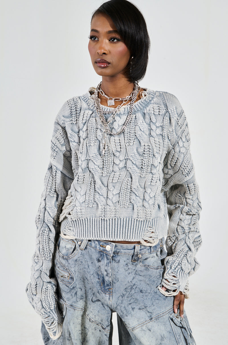 Against a plain backdrop, someone in distressed denim and the textured TALK TO ME NICE SWEATER, with layered necklaces, displays a calm expression as their right sleeve extends over their hand.