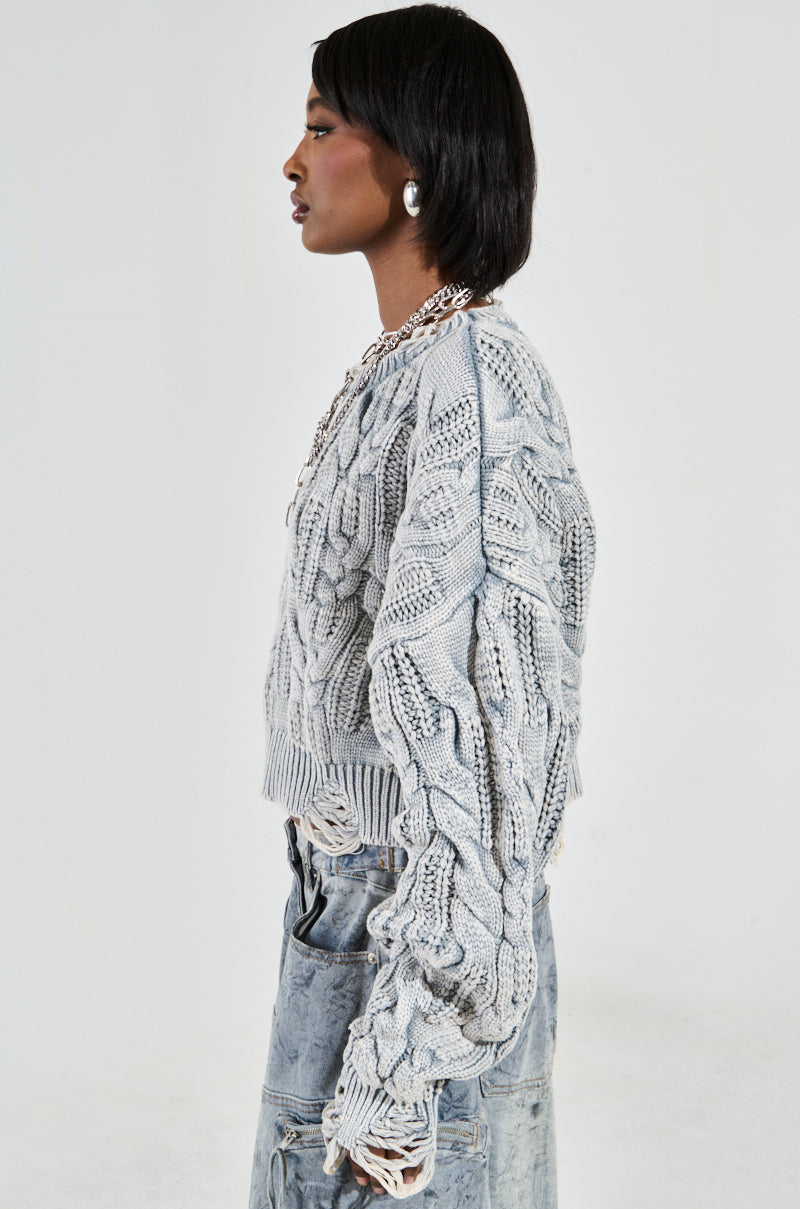A woman with short dark hair wears AKIRA Label's TALK TO ME NICE Sweater, a chunky gray cable knit style, paired with a long light blue denim skirt. Large silver hoop earrings and a silver chain necklace complete her profile against a plain white backdrop.