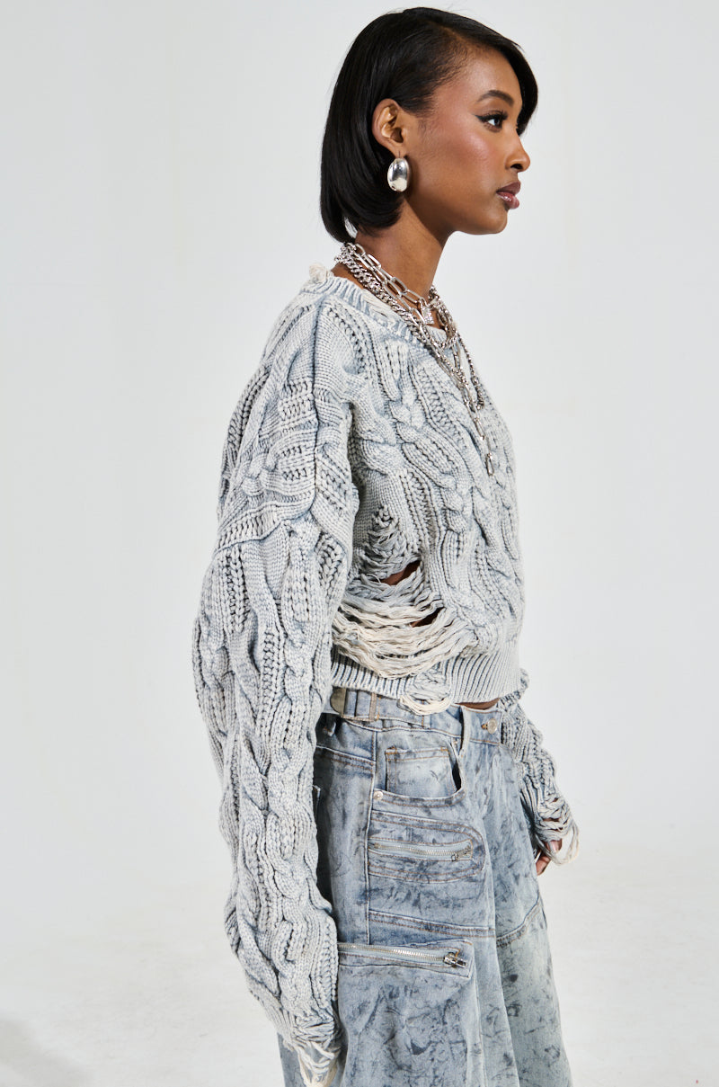 A person stands in profile against a light backdrop, wearing the TALK TO ME NICE SWEATER with distressed detailing and light blue, worn-look jeans. They have short hair and accessorize with large hoop earrings and layered necklaces.