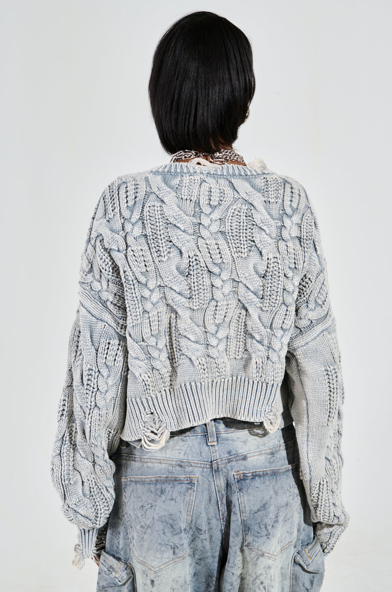 A person with short hair wears the grey chunky TALK TO ME NICE SWEATER with an oversized fit and dropped shoulders, paired with distressed blue jeans. The plain white background highlights the textured detailing of the outfit.