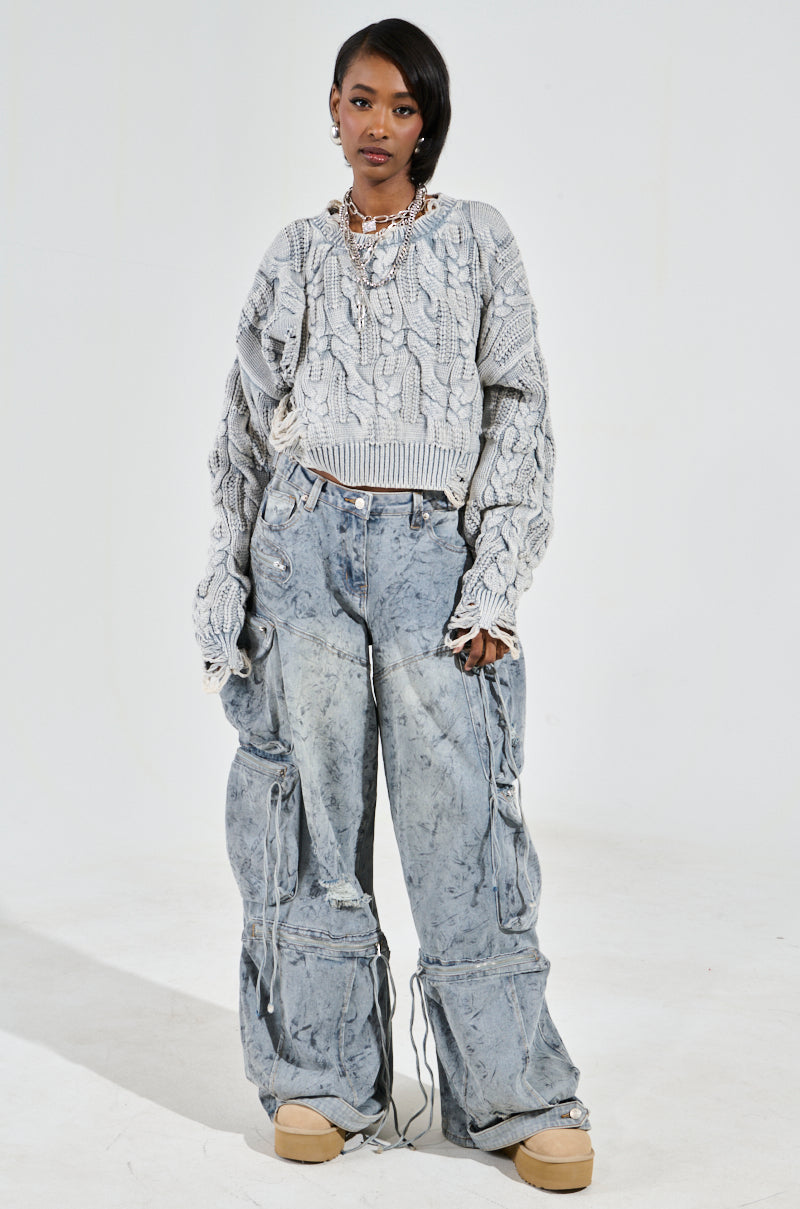 A person wears the AKIRA Label's TALK TO ME NICE sweater, an oversized gray cable knit style, with loose-fitting blue jeans featuring distressed details. They have short dark hair and a necklace against a light, plain background.