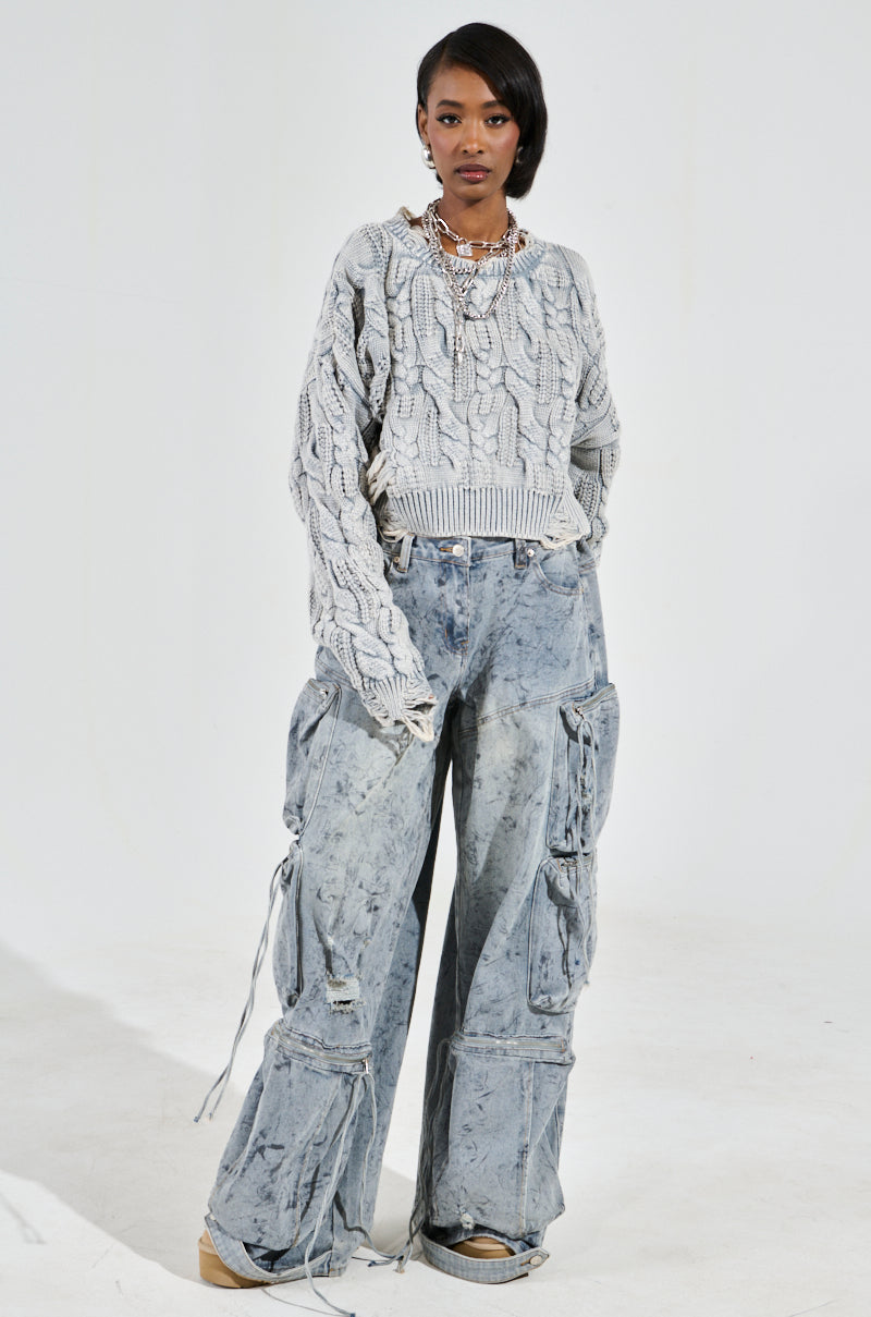 A person poses in a TALK TO ME NICE SWEATER, paired with wide-legged acid-wash cargo jeans with distressed detailing and multiple drawstring pockets. The look is completed with brown shoes and layered necklaces against a plain white background.