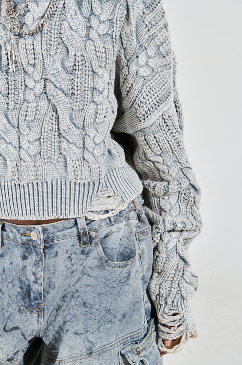 Someone wears an AKIRA Label TALK TO ME NICE gray cable-knit sweater with distressed detailing, teamed with faded acid-wash jeans, creating a casual, rugged look focused on texture.