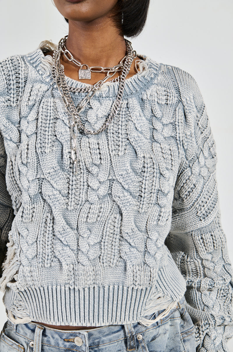 A person wearing a TALK TO ME NICE SWEATER by AKIRA Label, light gray with a cable-knit textured pattern, and layered silver chains stands against a plain background. The sweater pairs perfectly with light blue jeans, creating a stylish look.