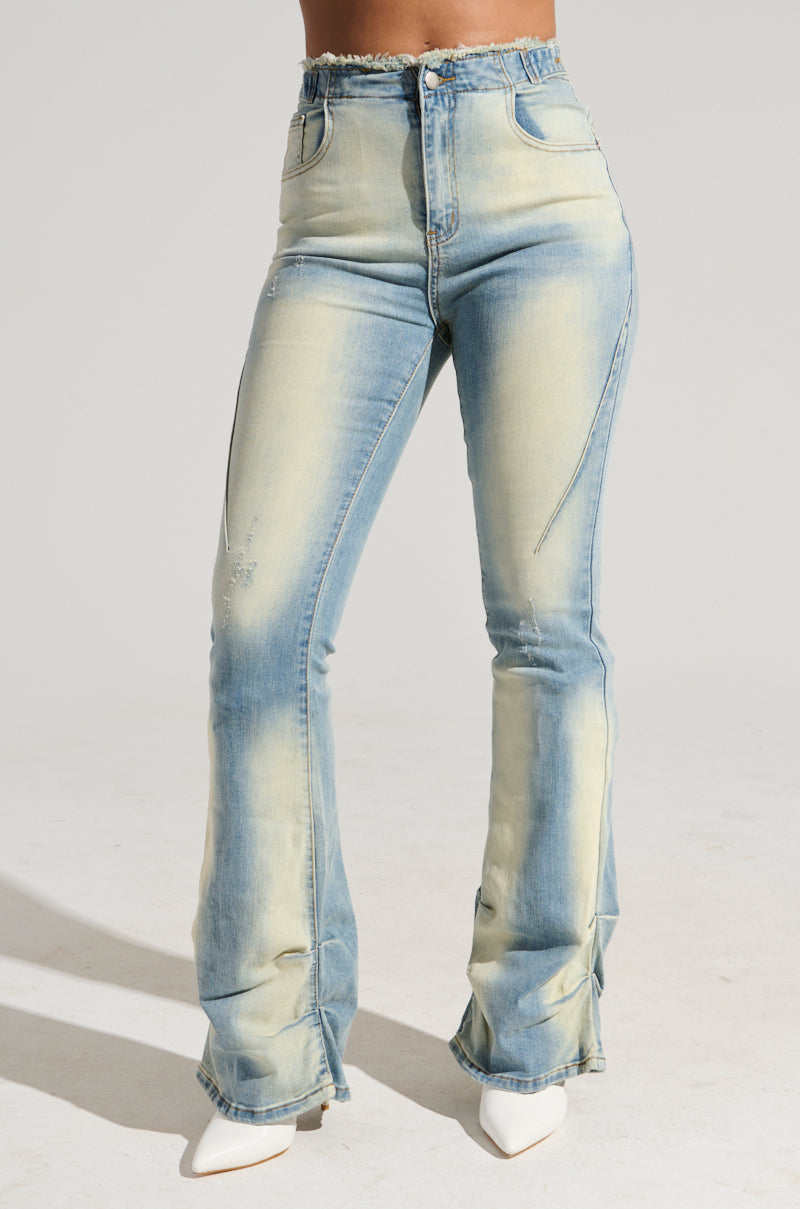 The model is standing and showcasing the WASHED UP FOR GOOD DENIM PANT, featuring a super stretchy, high-rise fit with light wash, slight distressing, front seam details, and flared legs. She is pairing them with white heeled shoes against a plain, light-colored background.