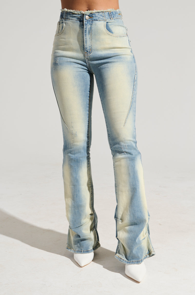 A person is wearing the "Washed Up For Good" denim pants, featuring light blue high-rise flared jeans with distressed details and frayed hems. They are also sporting white pointed-toe shoes against a plain, light-colored background.