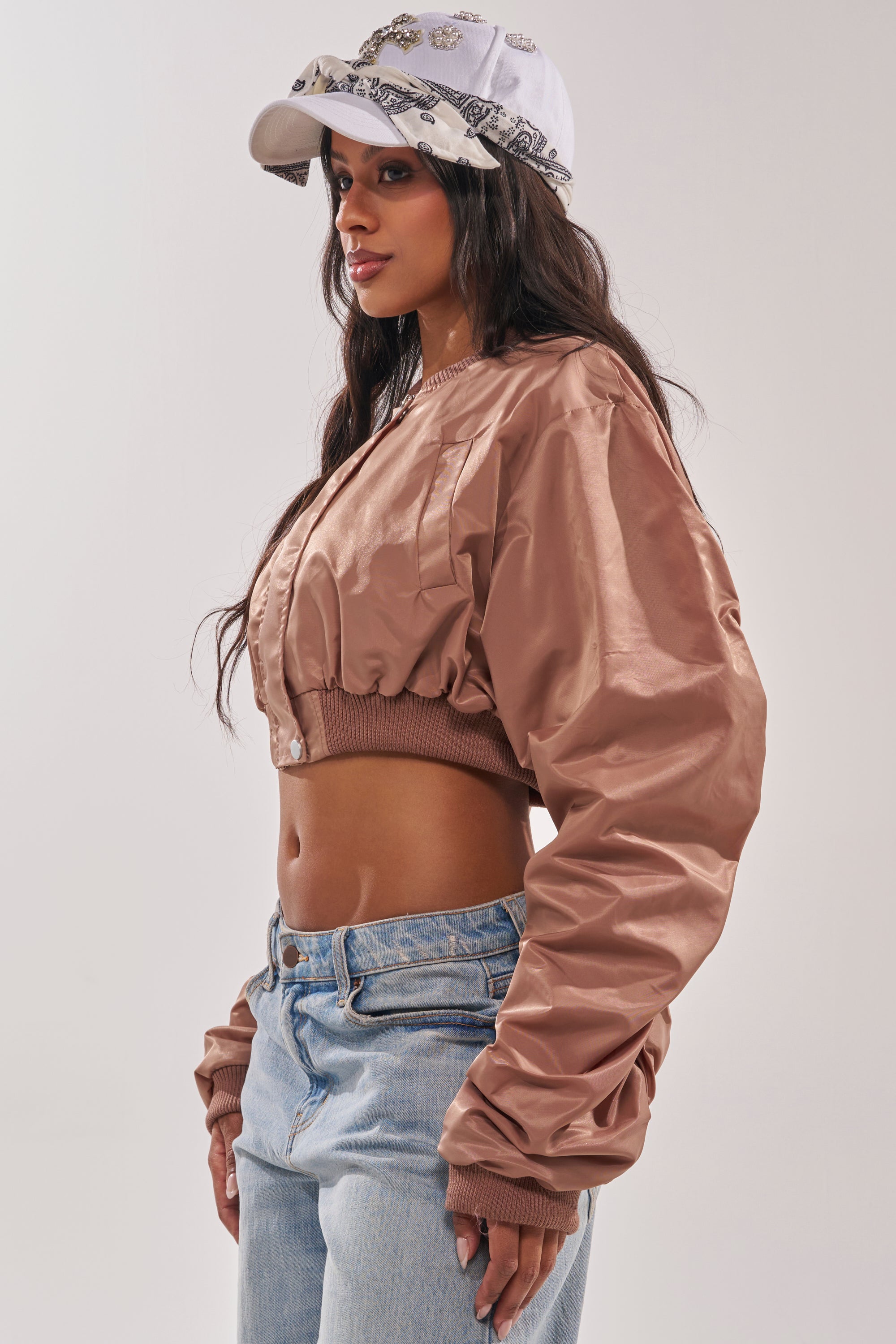 A woman in the TAKE IT EASY CROPPED BOMBER IN MAUVE, light blue jeans, and a white baseball cap with a patterned bandana stands sideways against a plain neutral background. 4