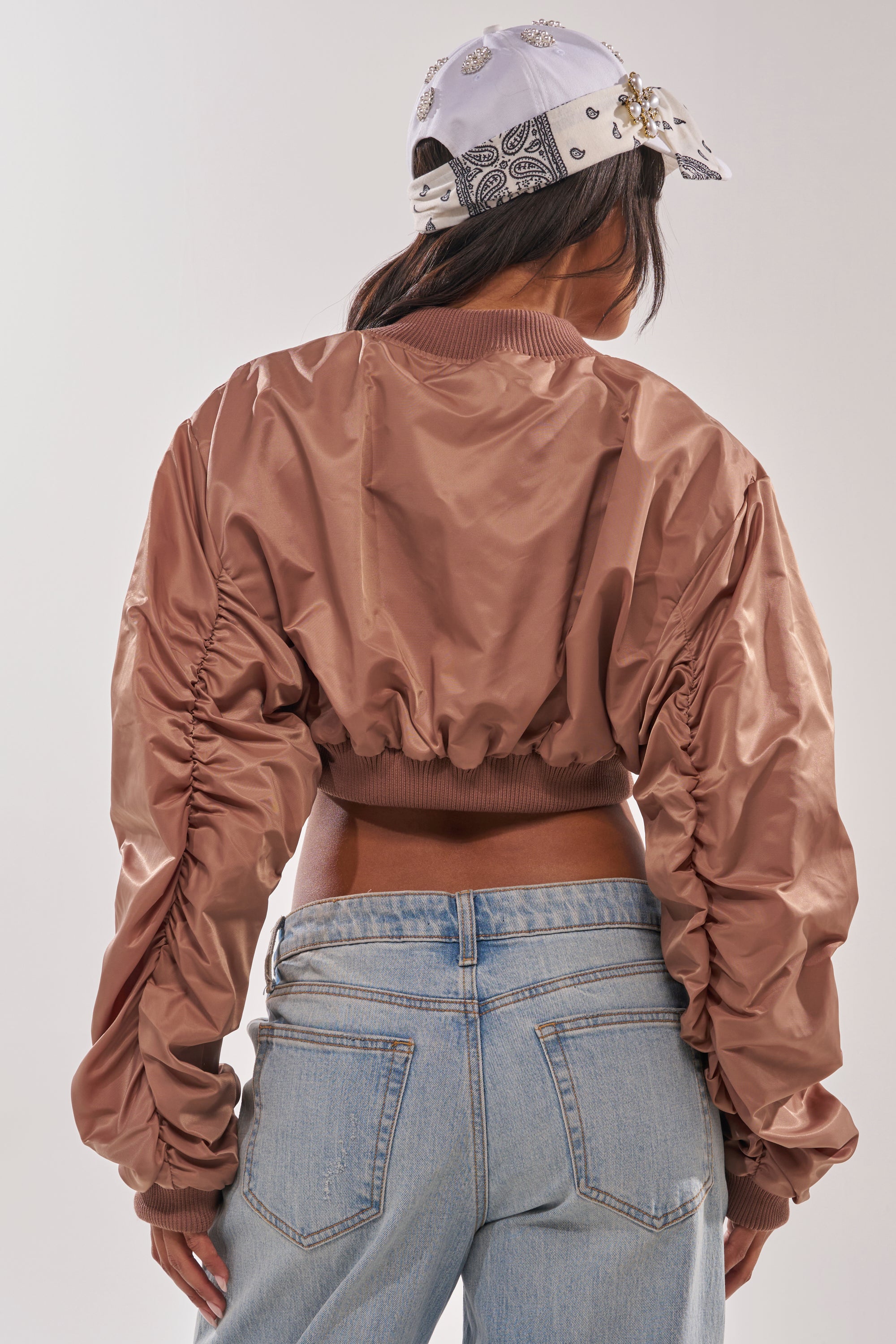 Someone is shown from the back wearing the TAKE IT EASY CROPPED BOMBER IN MAUVE, styled with ruched sleeves, paired with light blue jeans and a patterned light-colored baseball cap. 5