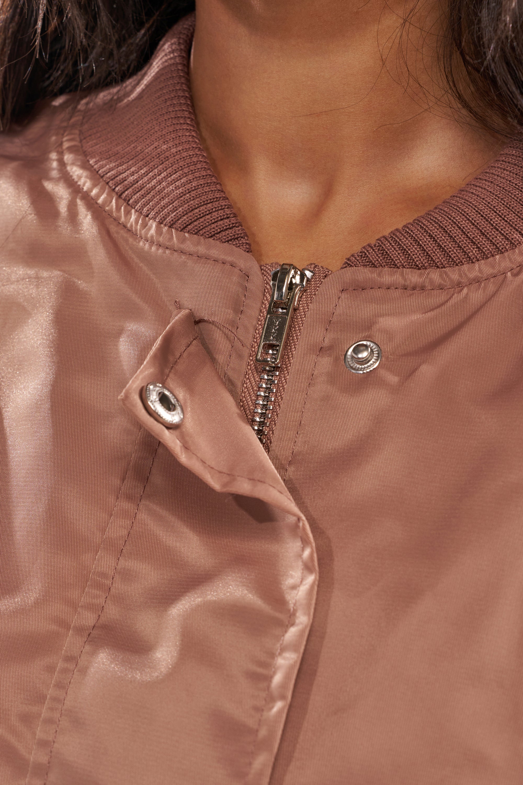 Close-up of a person wearing the TAKE IT EASY CROPPED BOMBER IN MAUVE, a shiny light brown women's jacket with zipper and snap button closure, highlighting the collar and upper chest area. 8
