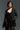 A woman in a chic black lace ensemble confidently poses against a dark backdrop. She is wearing the EASY DOES IT RELAXED FIT BLAZER along with lace gloves and a gold bracelet, complemented by dramatic earrings. Her hair is short and styled elegantly.