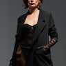 A woman in a chic black lace ensemble confidently poses against a dark backdrop. She is wearing the EASY DOES IT RELAXED FIT BLAZER along with lace gloves and a gold bracelet, complemented by dramatic earrings. Her hair is short and styled elegantly.