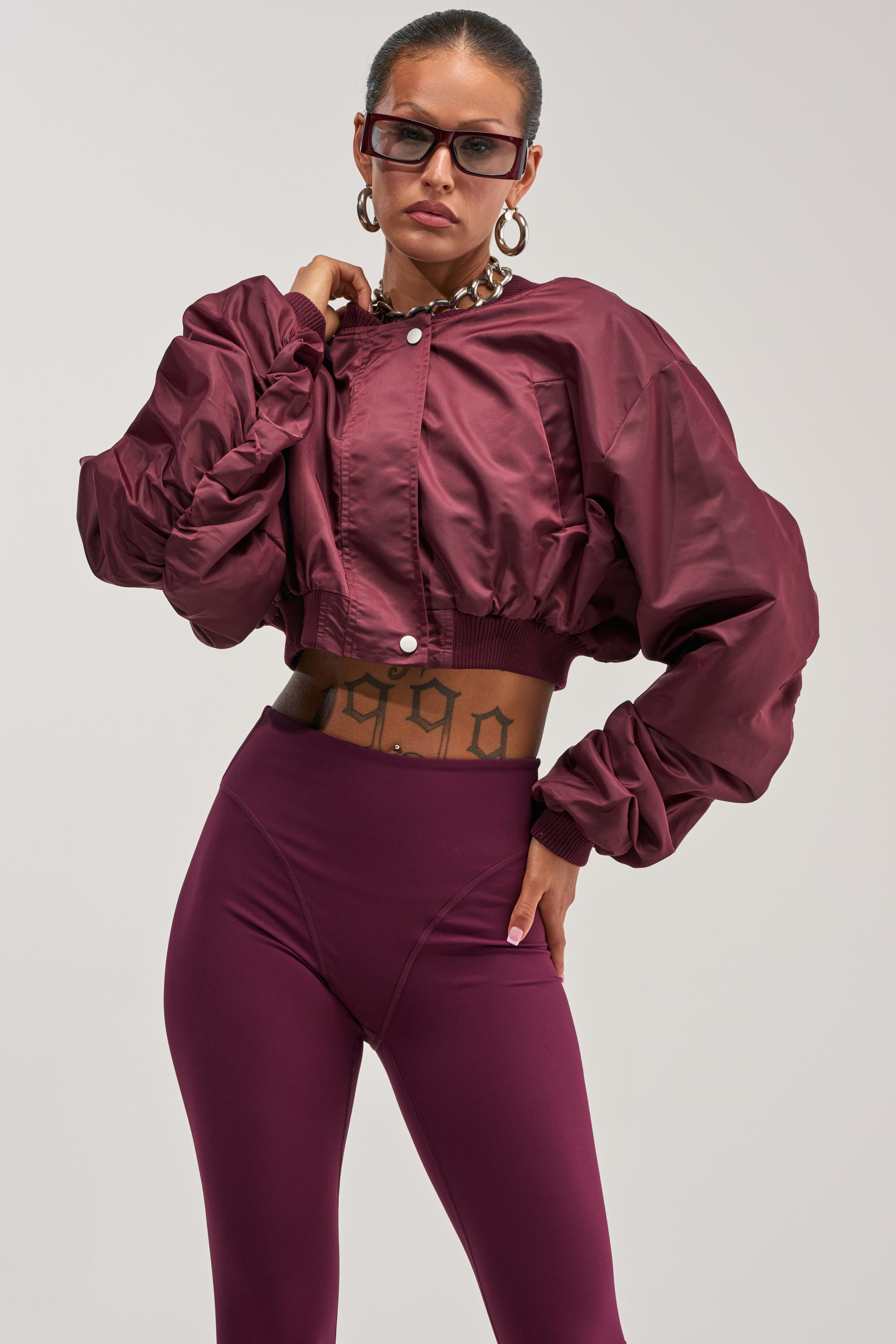 A woman poses confidently against a plain background wearing the TAKE IT EASY CROPPED BOMBER IN BURGUNDY paired with matching high-waisted leggings, accessorized with oversized glasses, a chunky necklace, and showcasing a visible "1990" tattoo on her torso.
