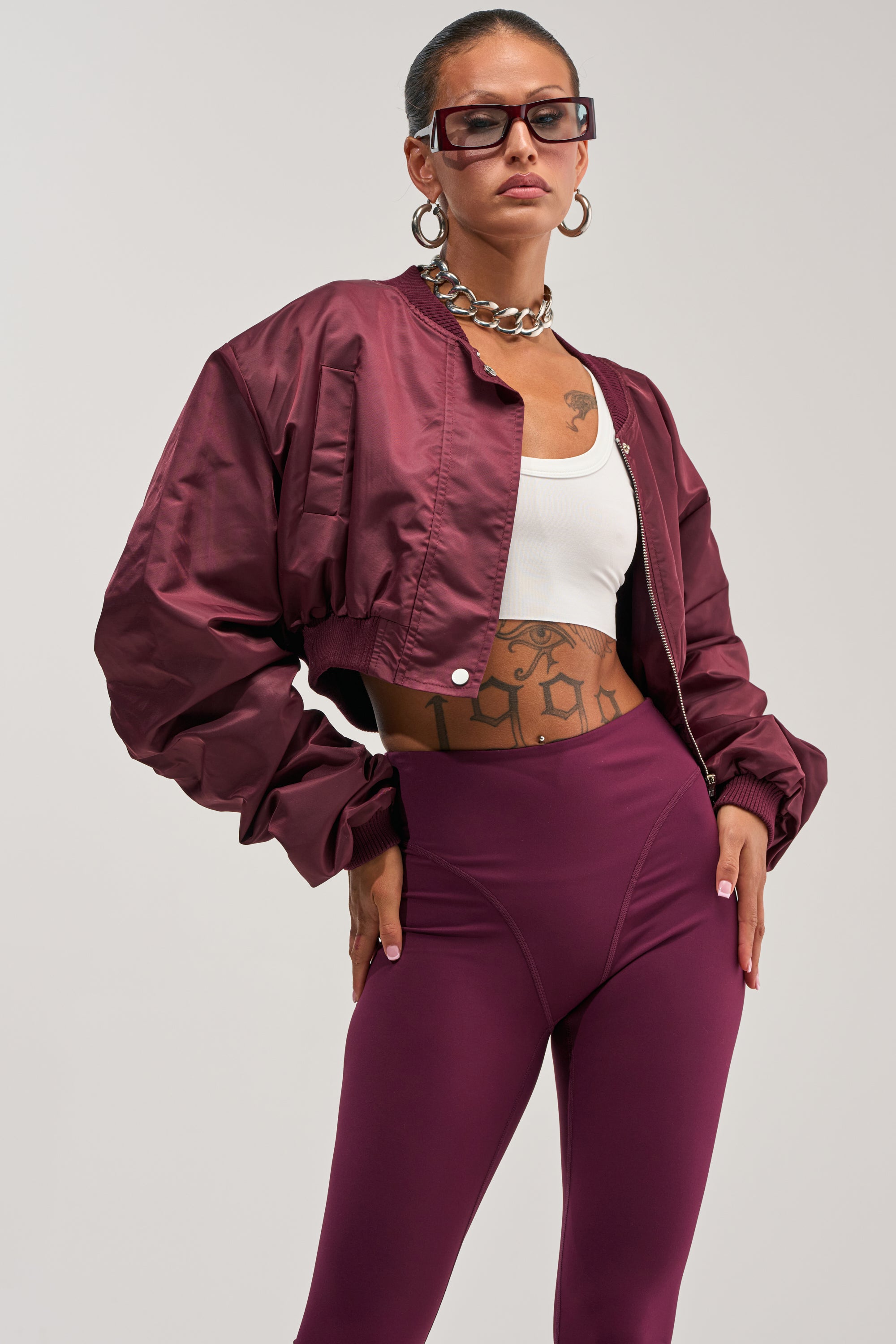 Clad in the TAKE IT EASY CROPPED BOMBER IN BURGUNDY, a person strikes a confident pose. They style the jacket with a white crop top and coordinating maroon leggings, finishing the look with oversized sunglasses, hoop earrings, a chunky chain necklace, and visible torso tattoos.