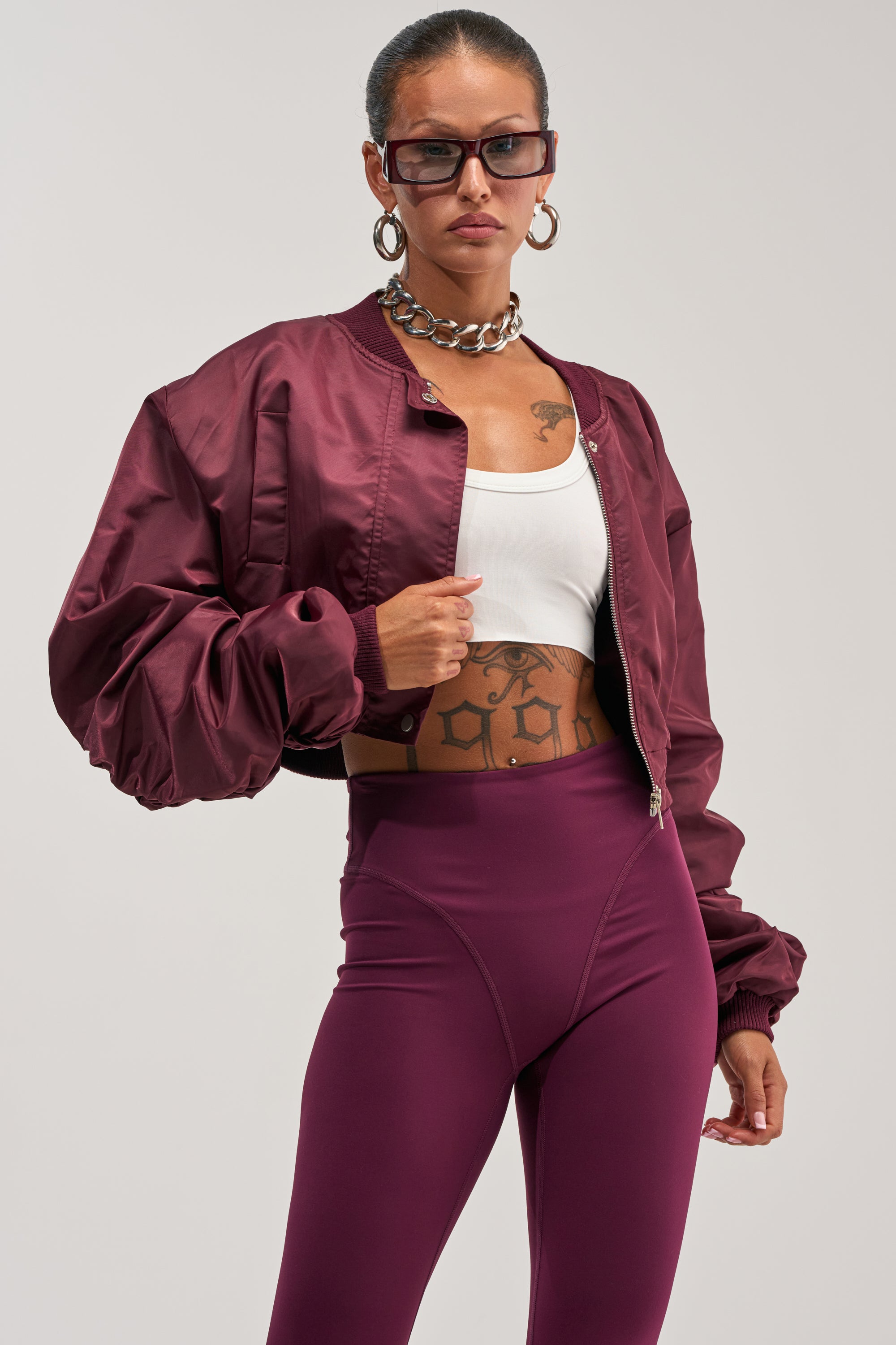 A woman poses confidently against a plain background, showcasing the TAKE IT EASY CROPPED BOMBER IN BURGUNDY with ruched sleeves, styled with matching high-waisted leggings, a white crop top, chunky necklace, large hoop earrings, and sunglasses.