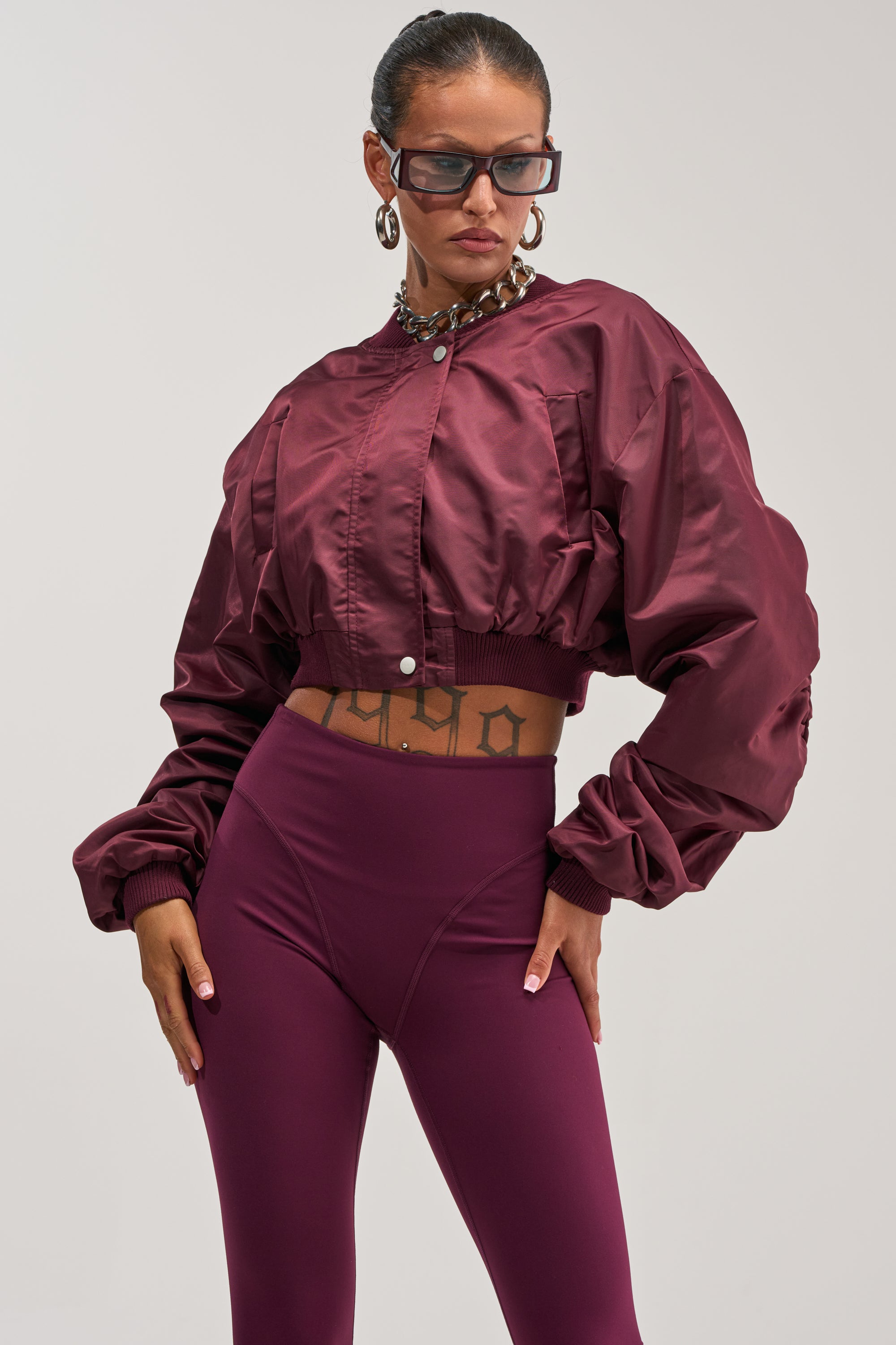 A woman models the TAKE IT EASY CROPPED BOMBER IN BURGUNDY with matching high-waisted leggings, styled with sunglasses, large hoop earrings, and a chunky necklace, posing confidently with her hands on her hips against a plain background.