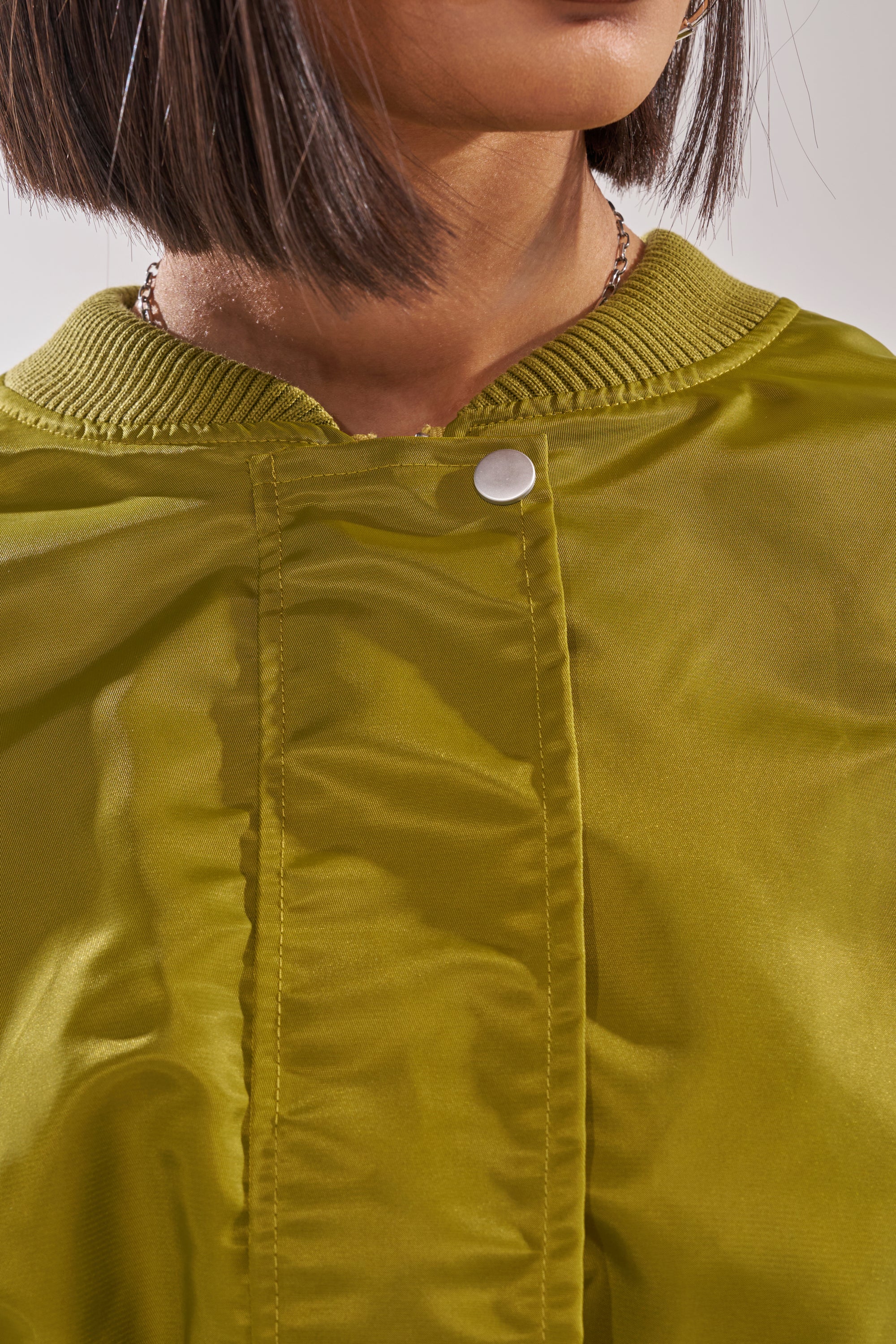A close-up shows a person with a bob haircut wearing the TAKE IT EASY CROPPED BOMBER IN GREEN, featuring an olive green hue, ribbed collar, and single white snap button. Only their lower face and neck are visible. 9