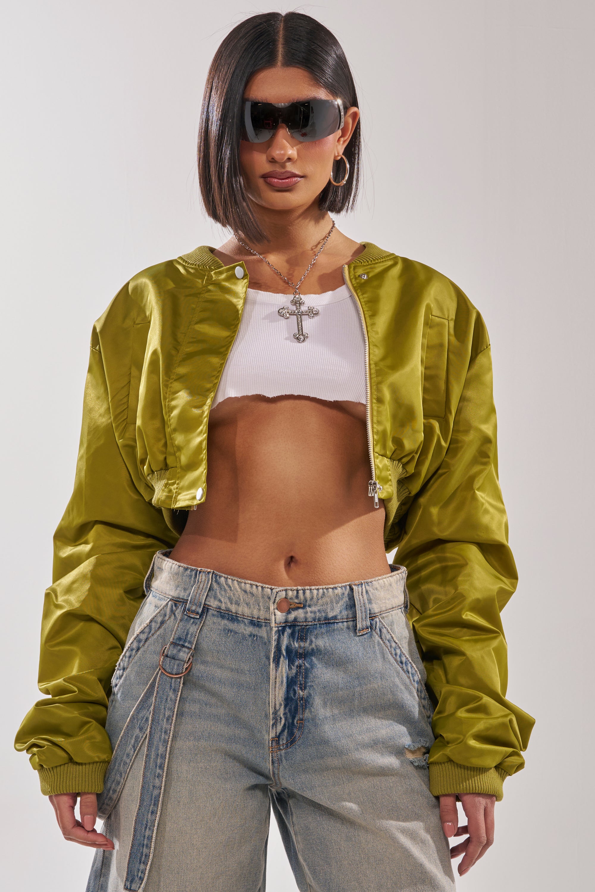 A woman with a sleek bob haircut wears dark sunglasses, the TAKE IT EASY CROPPED BOMBER IN GREEN, a white crop top, baggy blue jeans, and a silver cross necklace as she stands confidently against a plain background. 0