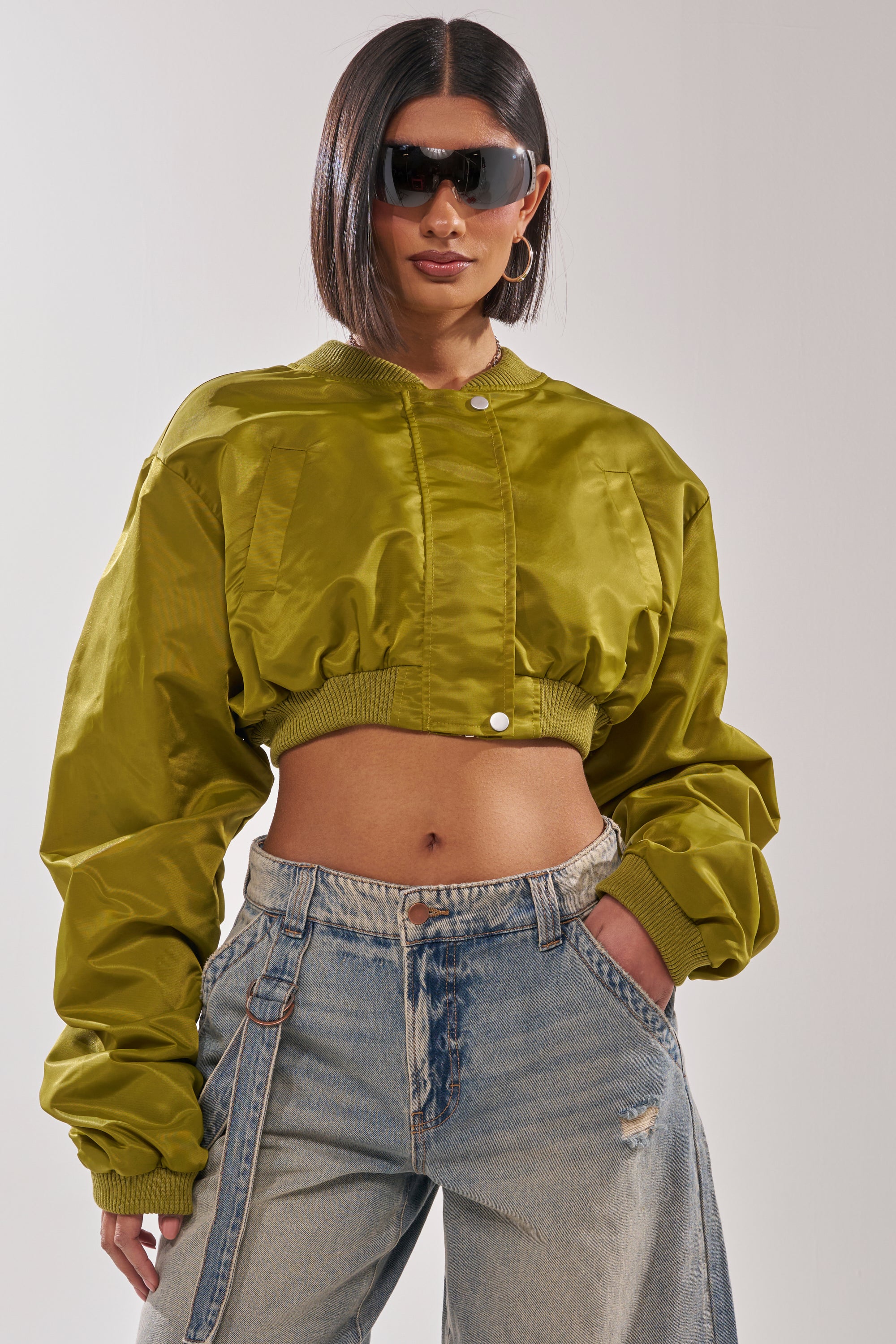 A woman wearing the TAKE IT EASY CROPPED BOMBER IN GREEN, paired with large black sunglasses, hoop earrings, and light blue high-waisted jeans stands with her hands in her pockets against a plain background. 1
