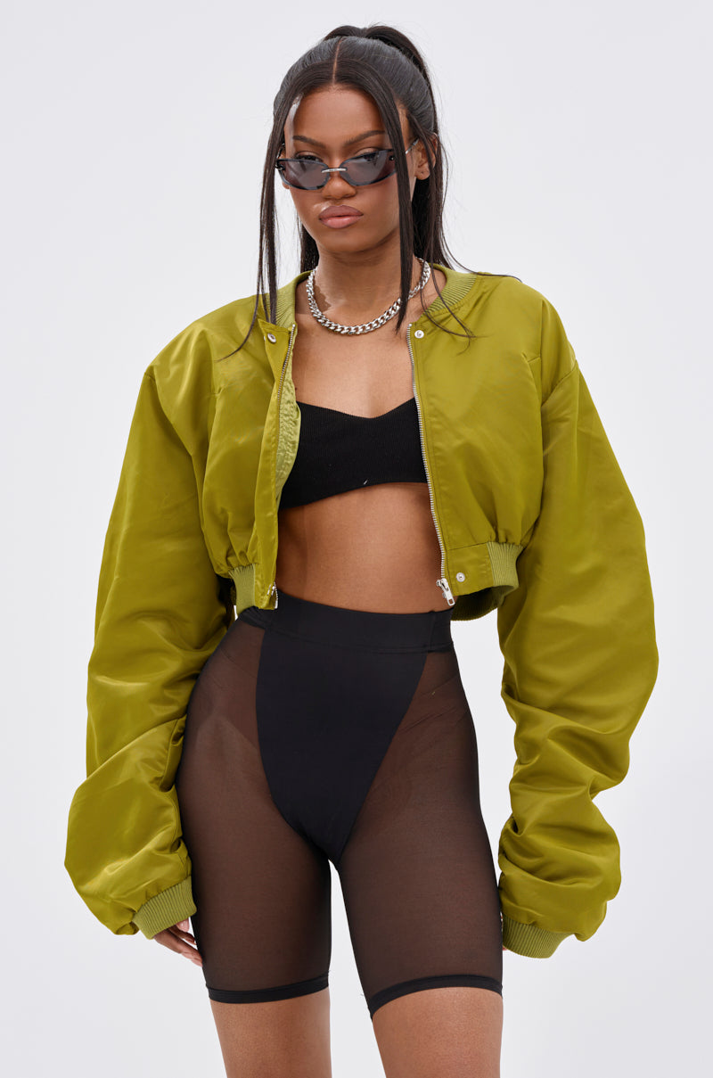 She wears the TAKE IT EASY CROPPED BOMBER IN GREEN over a black crop top and sheer black high-waisted shorts, accessorized with sunglasses, a chain necklace, and two ponytails, standing against a light background.