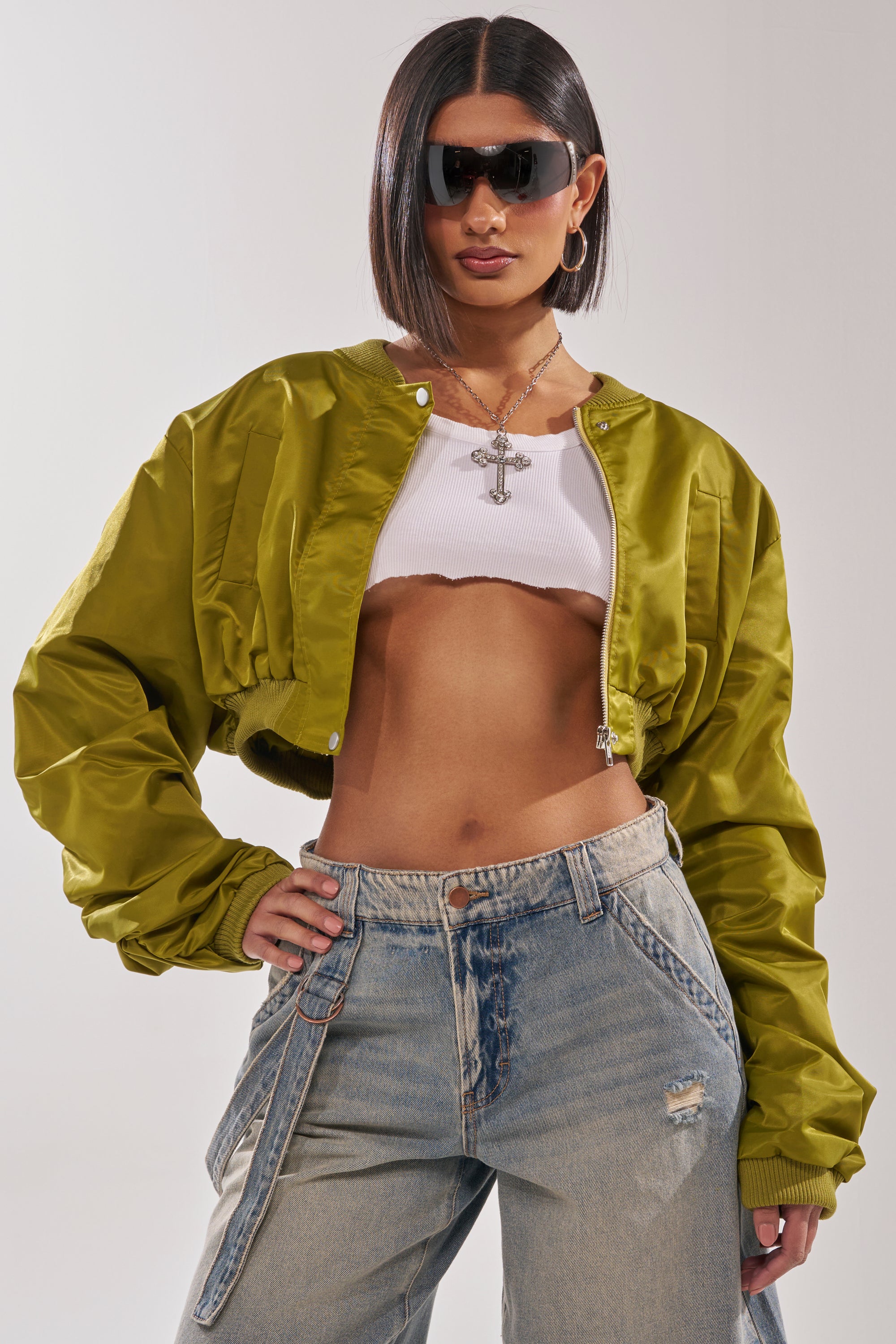 A woman wearing the TAKE IT EASY CROPPED BOMBER IN GREEN, large sunglasses, a white ribbed crop top, distressed jeans, hoop earrings, and a cross necklace poses confidently with one hand on her hip. 2