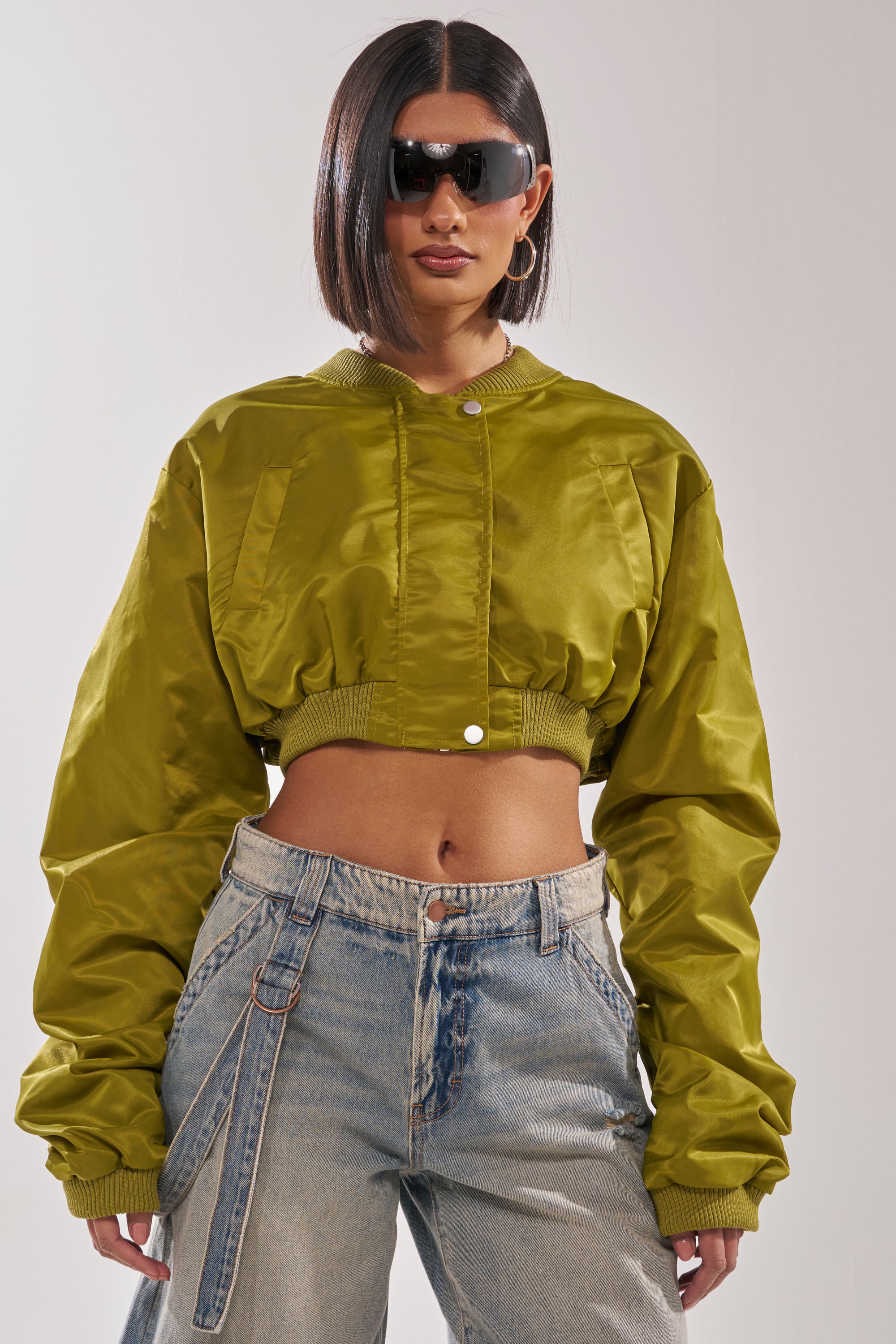 Wearing the TAKE IT EASY CROPPED BOMBER IN GREEN with loose light-wash belted jeans, this person sports short straight hair, large hoop earrings, and dark sunglasses. 3