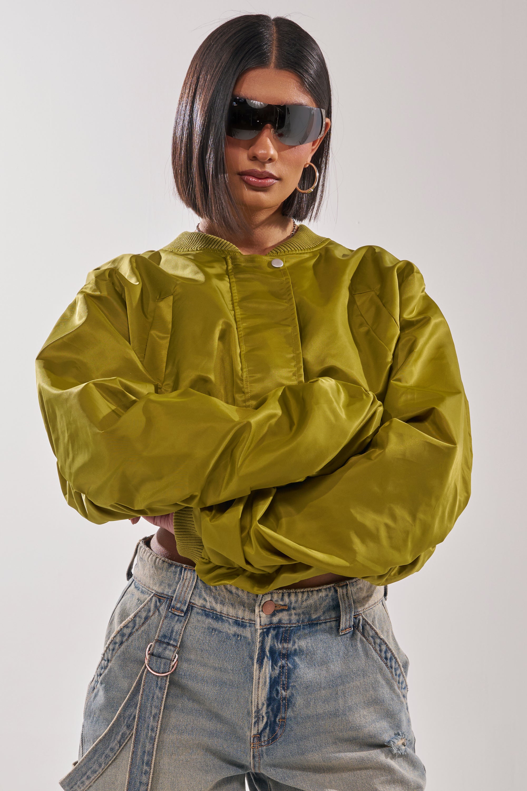 A woman with a sleek bob, wearing large black sunglasses, light-wash chain-detail jeans, and the TAKE IT EASY CROPPED BOMBER IN GREEN, stands with arms crossed against a plain background. 4