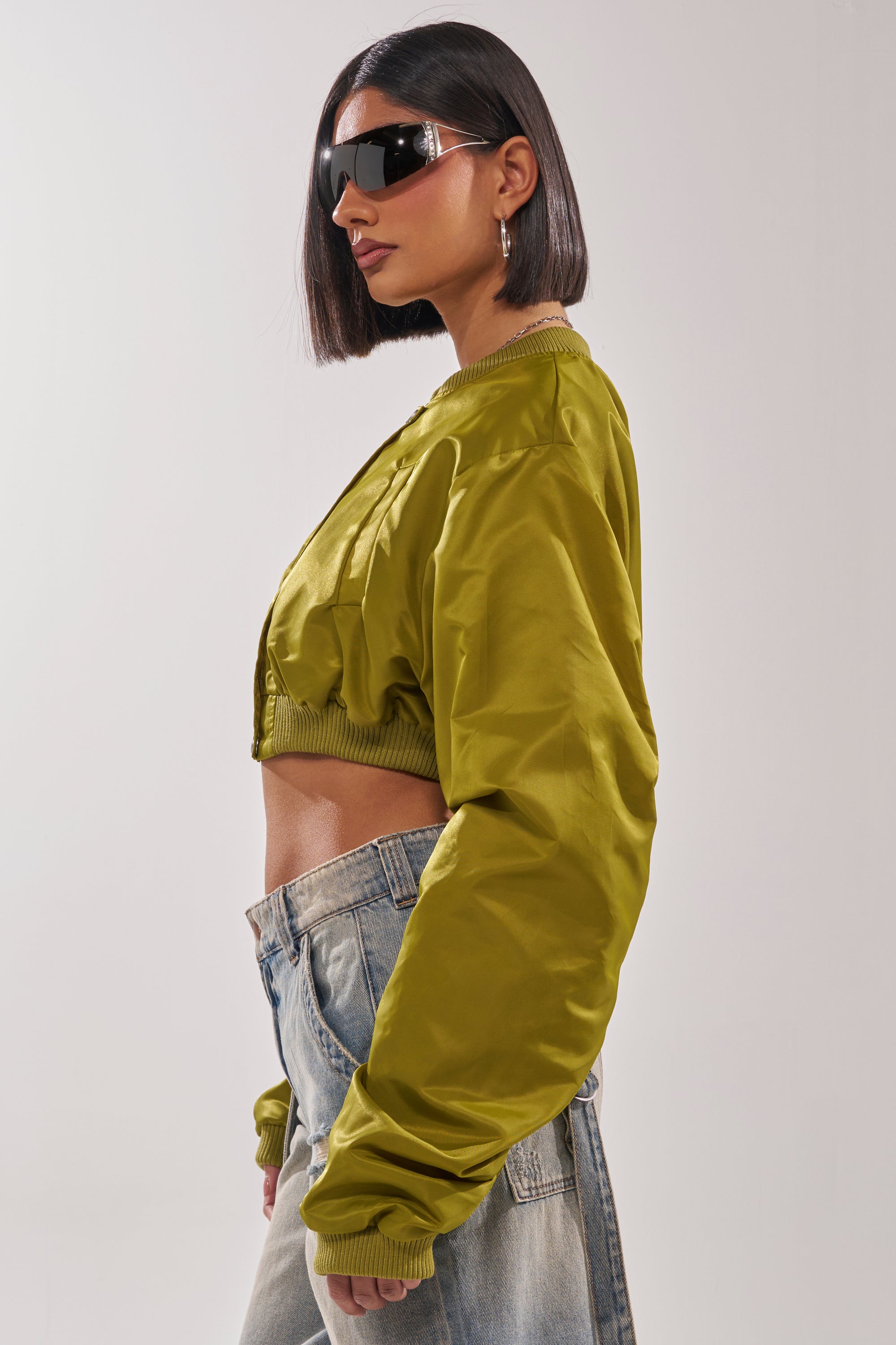 A woman with a sleek bob and large black sunglasses stands in profile, wearing light blue jeans and the TAKE IT EASY CROPPED BOMBER IN GREEN against a plain light background. 5