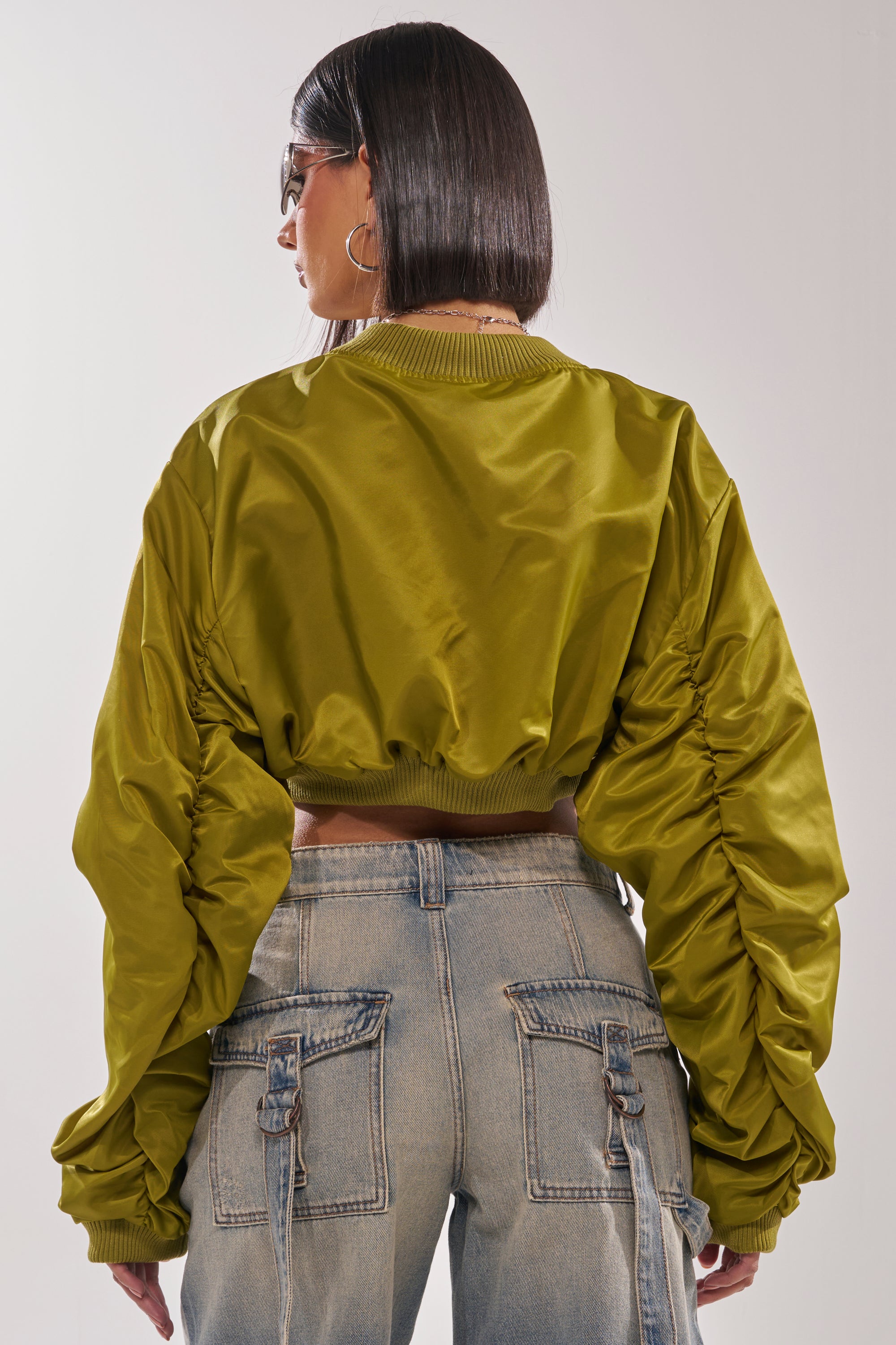 A woman with straight shoulder-length hair wears sunglasses, the TAKE IT EASY CROPPED BOMBER IN GREEN, and light blue cargo jeans, shown from the back against a plain background. 6
