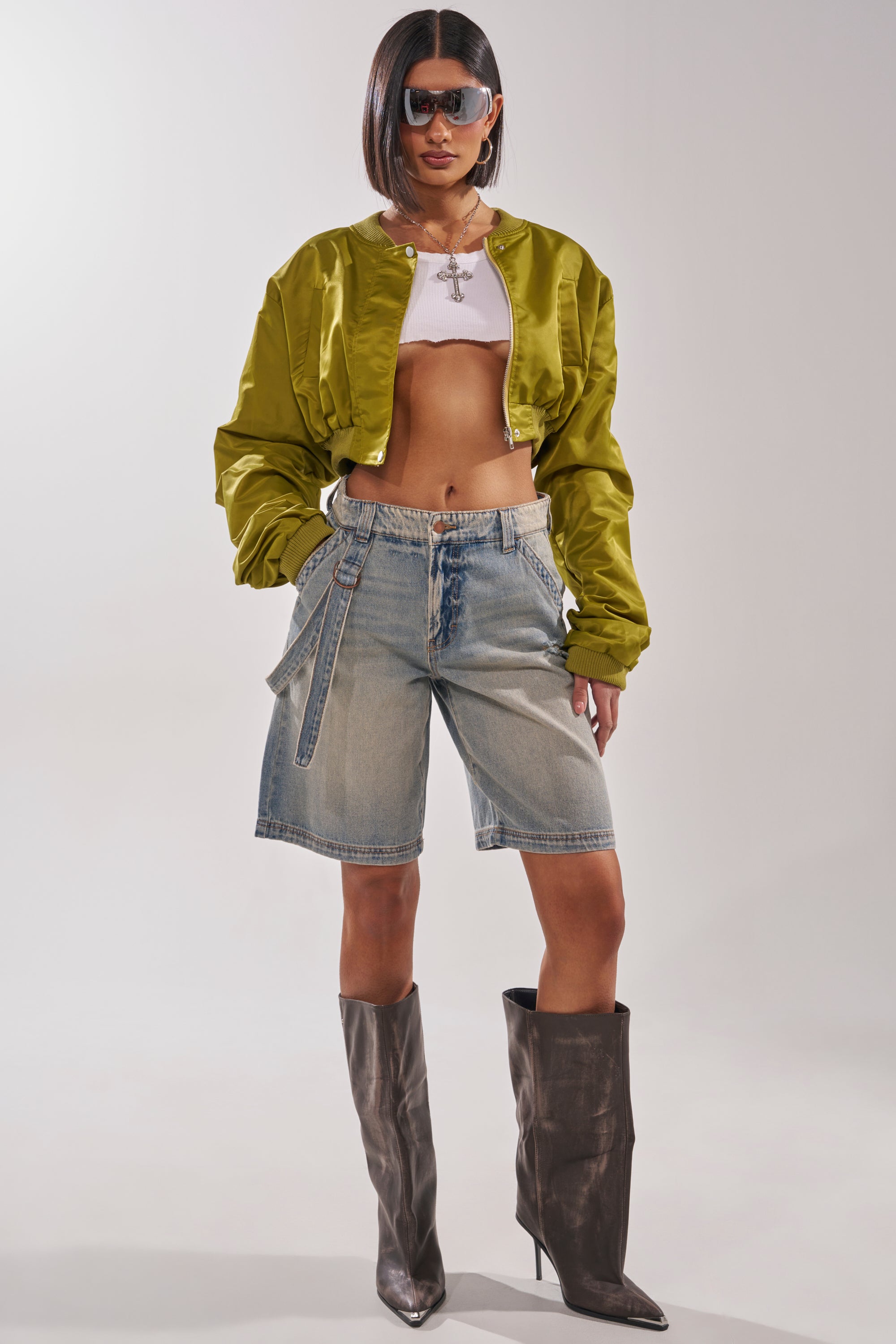 A woman poses confidently in the TAKE IT EASY CROPPED BOMBER IN GREEN, styled over a white crop top, denim Bermuda shorts, tall brown heeled boots, sunglasses, and layered necklaces against a plain background. 7
