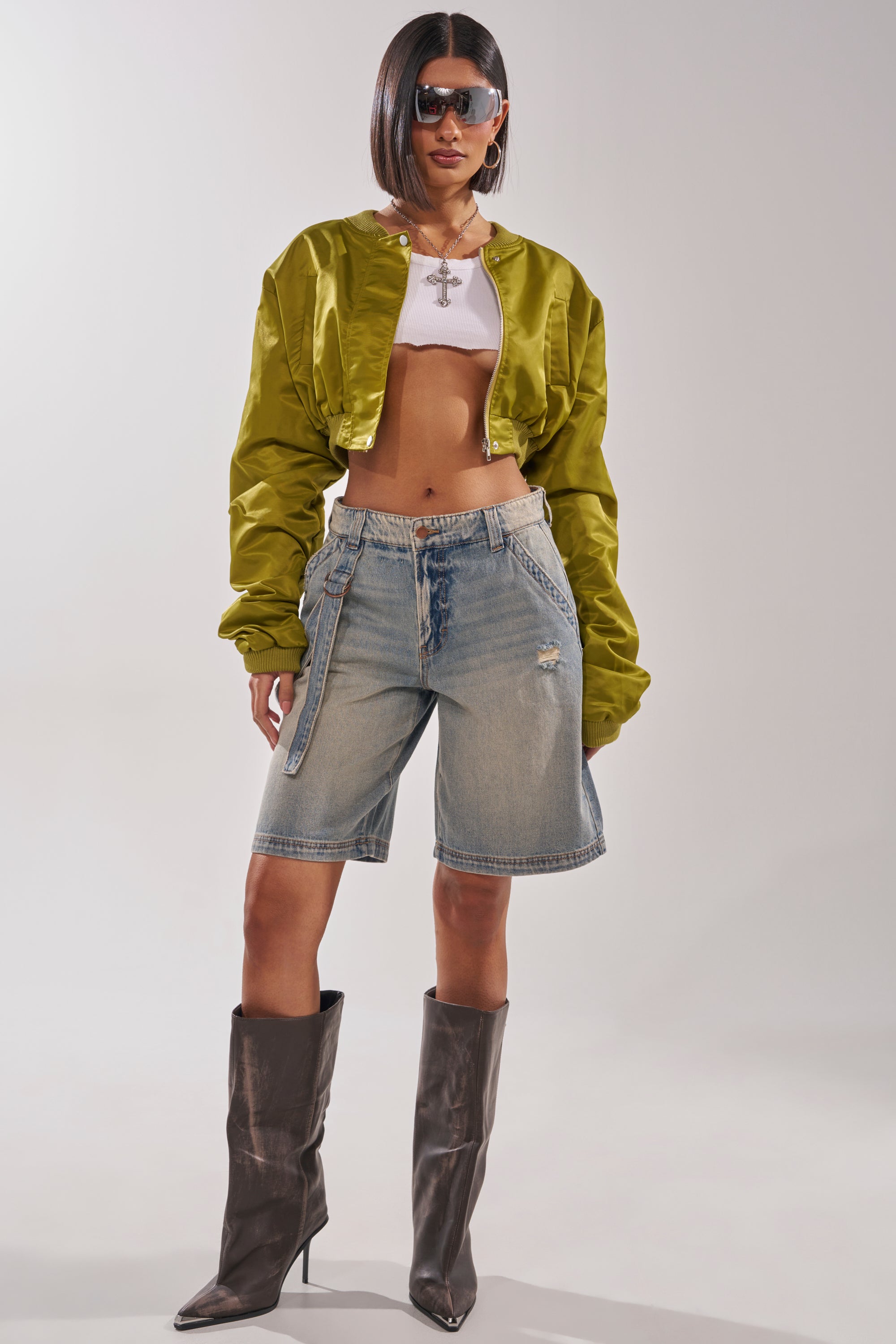 A woman poses confidently against a plain background in the TAKE IT EASY CROPPED BOMBER IN GREEN, paired with a white crop top, distressed denim Bermuda shorts, tall brown boots, sunglasses, and jewelry. 8