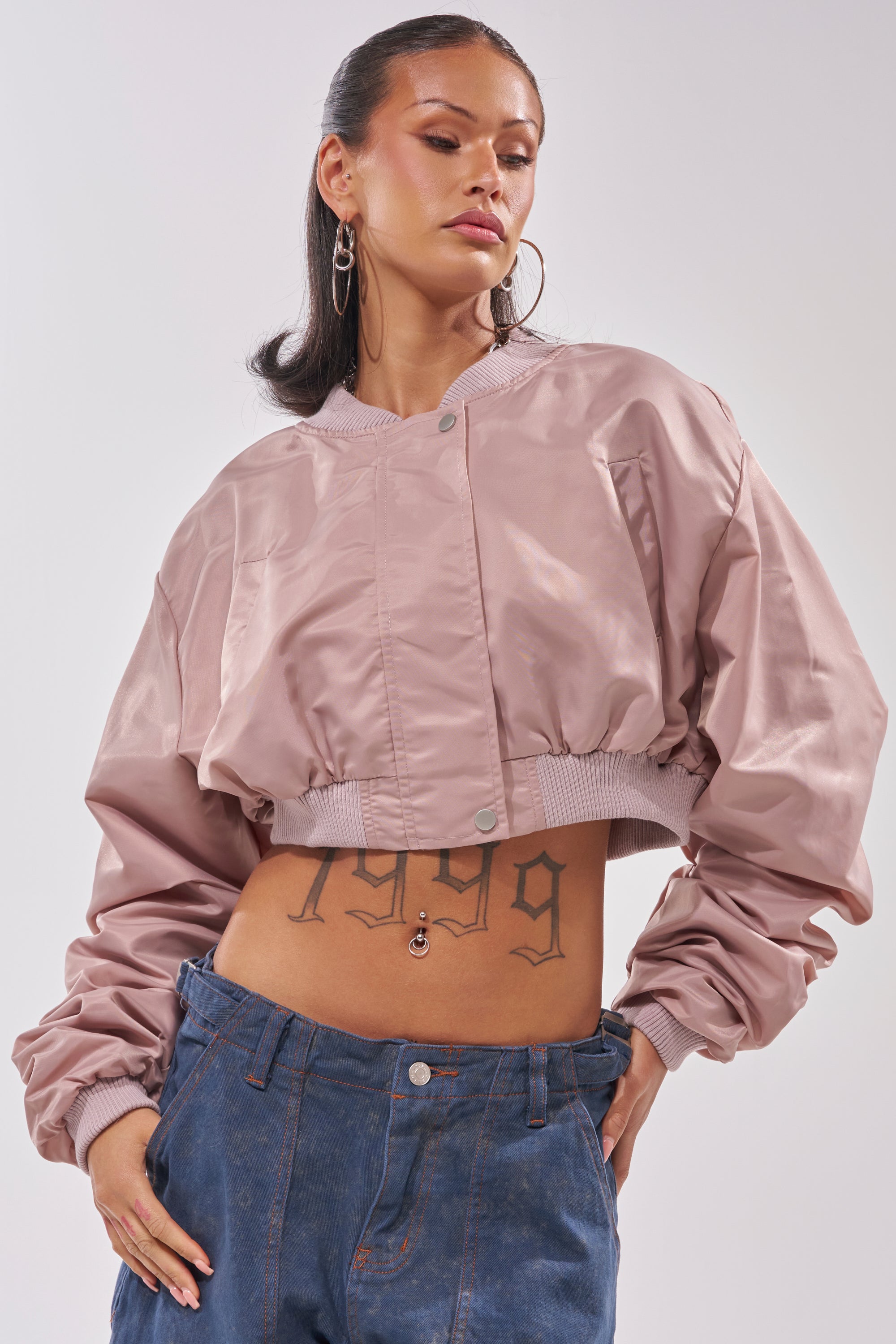 A woman models the TAKE IT EASY CROPPED BOMBER IN MAUVE with blue jeans, accessorized with hoop earrings, a belly button piercing, and a "1999" tattoo on her lower stomach. One hand is casually tucked in her pocket. 0