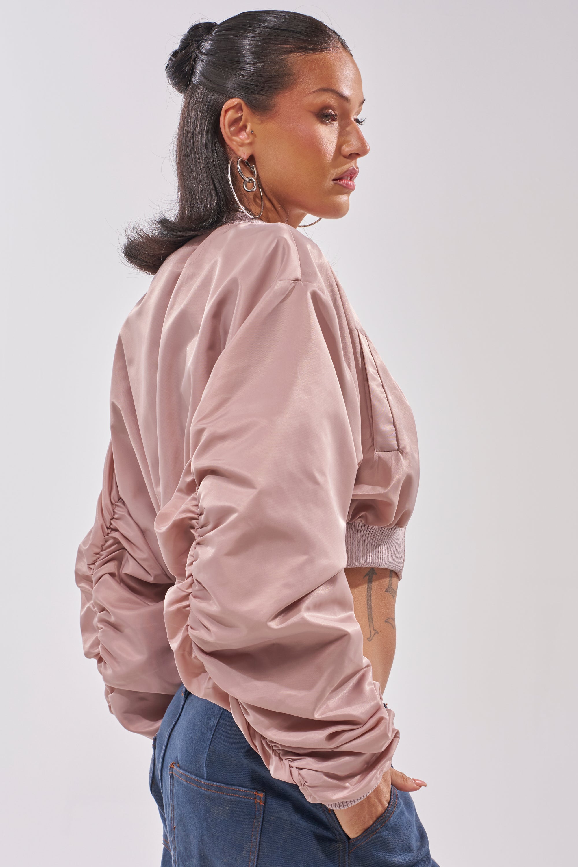 A woman with long, dark hair styled back wears the TAKE IT EASY CROPPED BOMBER IN MAUVE and blue jeans. She stands sideways, looking away from the camera with one hand in her pocket. 2