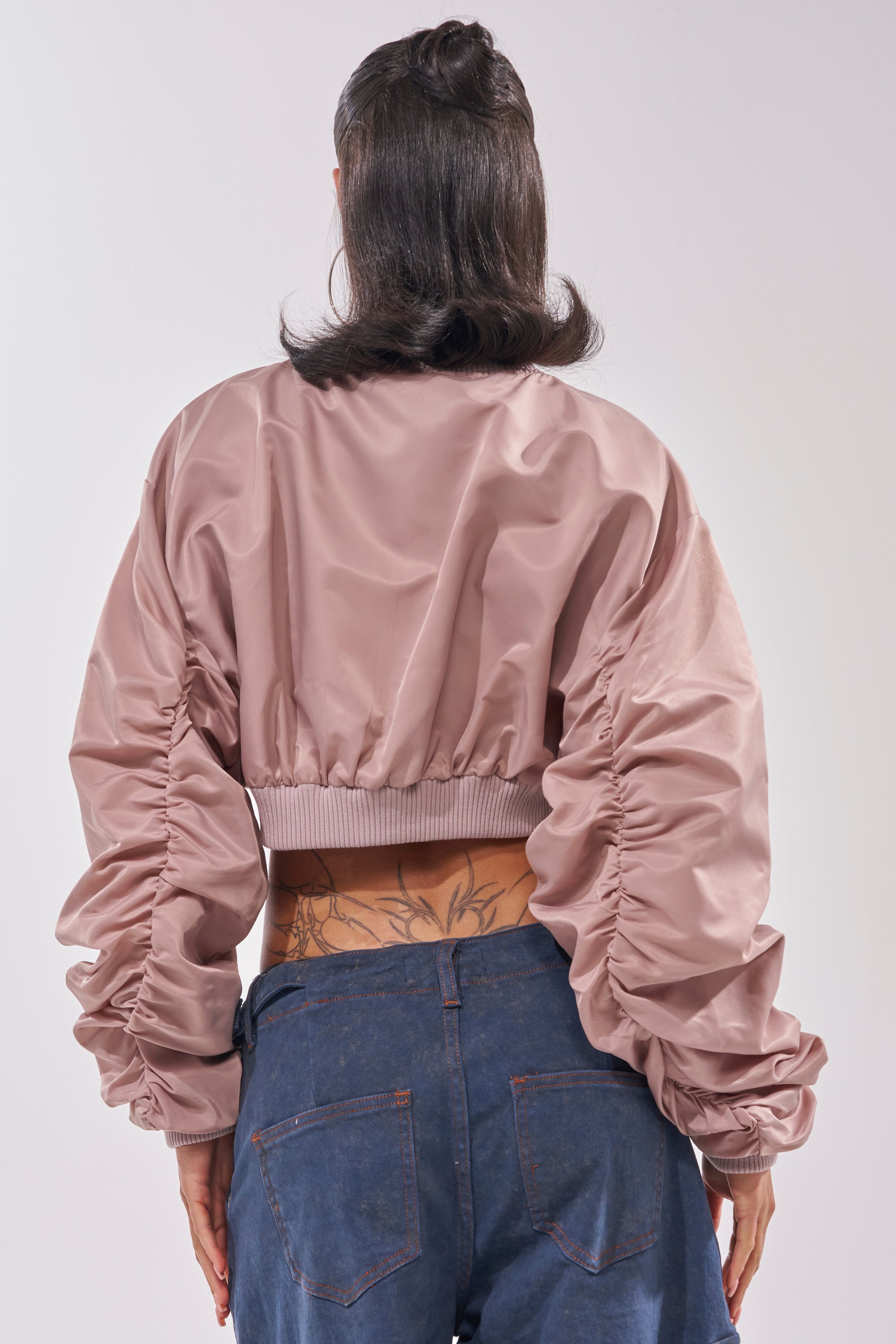 A person with medium-length dark hair is seen from behind wearing the TAKE IT EASY CROPPED BOMBER IN MAUVE and blue jeans, revealing a floral tattoo on their lower back. 3