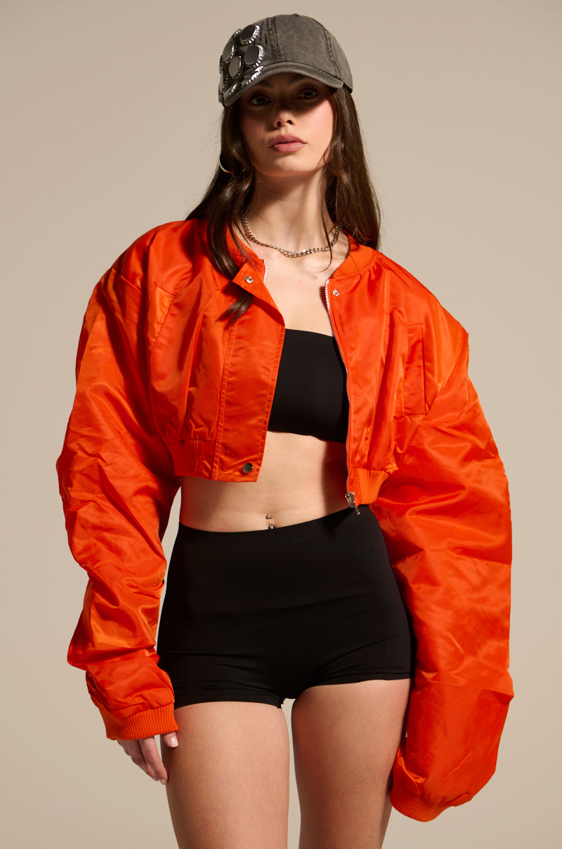 A woman wearing the TAKE IT EASY CROPPED BOMBER IN ORANGE, a black tube top, and black shorts stands against a beige background. She accessorizes with a gray cap and chain necklace, her hand resting by her side.