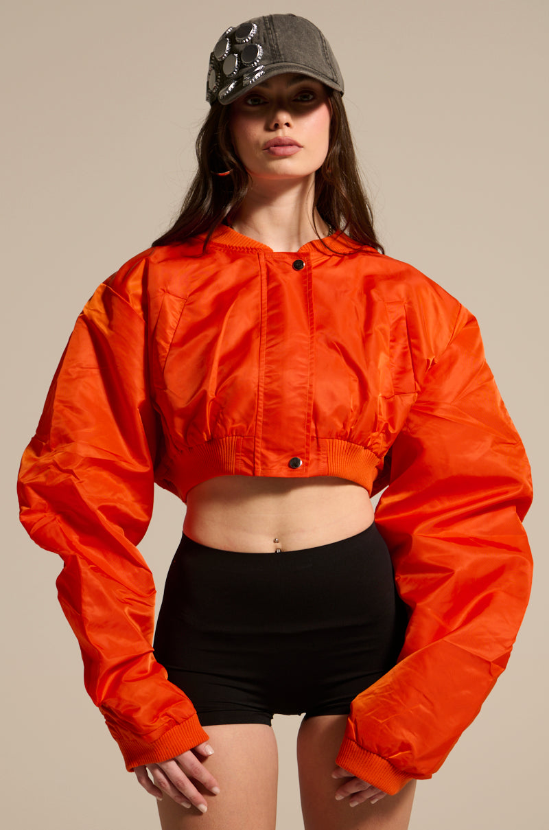 A woman in a gray embellished cap, the TAKE IT EASY CROPPED BOMBER IN ORANGE, and high-waisted black shorts stands against a neutral background.