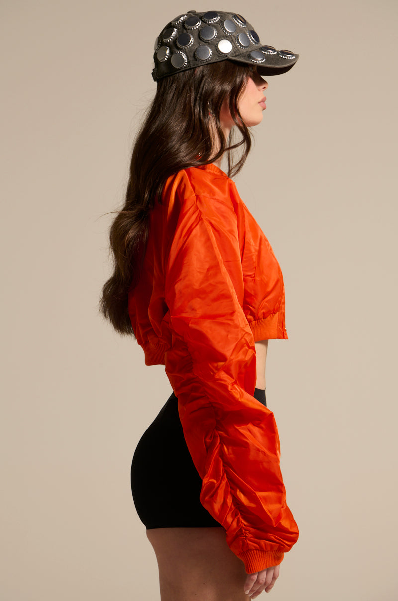 A woman stands in profile wearing the TAKE IT EASY CROPPED BOMBER IN ORANGE, paired with black shorts and a black baseball cap with large metallic circles, set against a plain beige background.