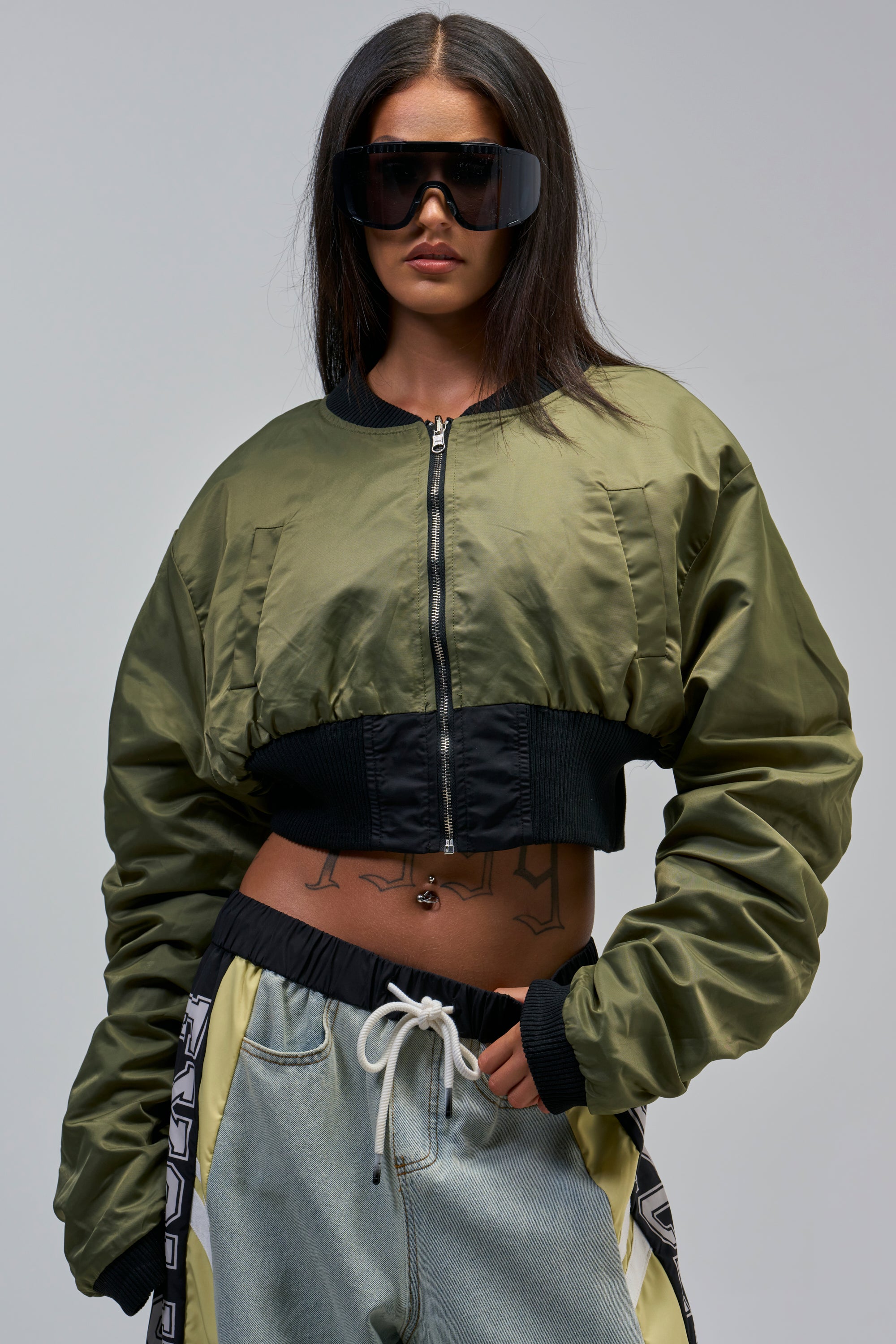 Woman with long dark hair wears the TAKE IT EASY REVERSIBLE BOMBER in olive green, paired with drawstring blue jeans. Hands in pockets, her stomach tattoos are on display.