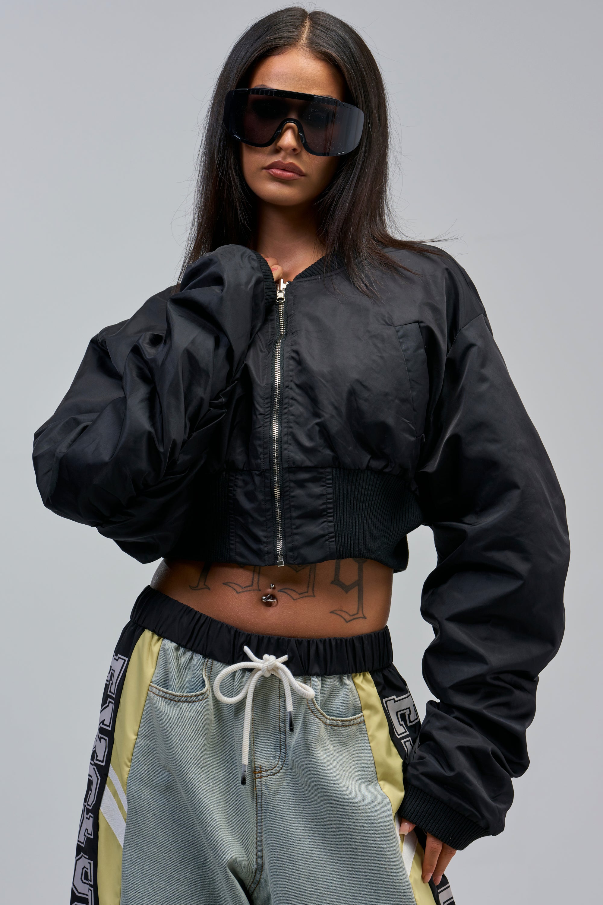 A woman in oversized sunglasses and the TAKE IT EASY REVERSIBLE BOMBER with loose drawstring pants poses confidently, showcasing streetwear style with long dark hair and tattoo lettering on her stomach.