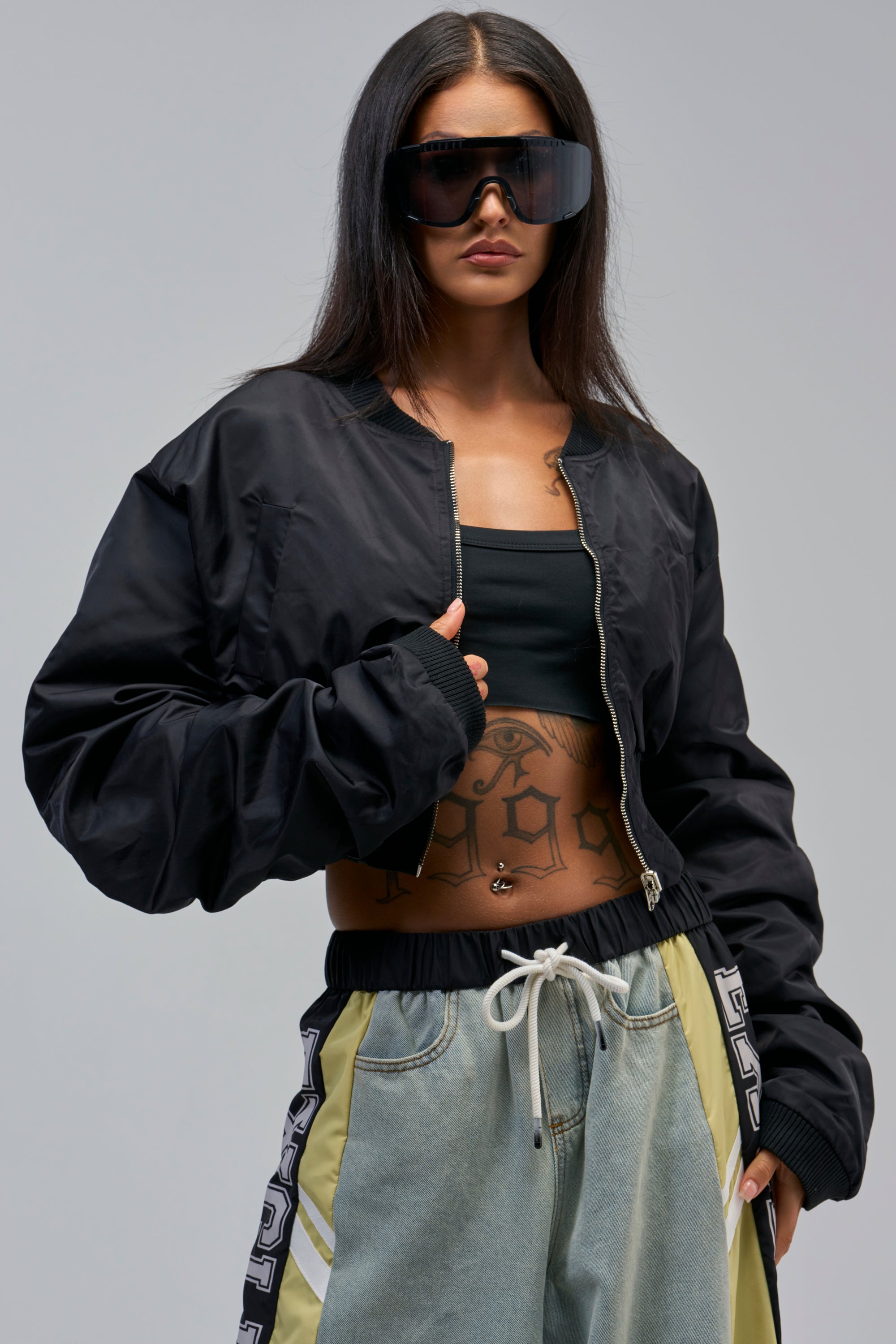 Woman wearing streetwear: oversized sunglasses, a black crop top, loose drawstring denim pants, and the TAKE IT EASY REVERSIBLE BOMBER. She has long dark hair and visible tattoos on her torso.